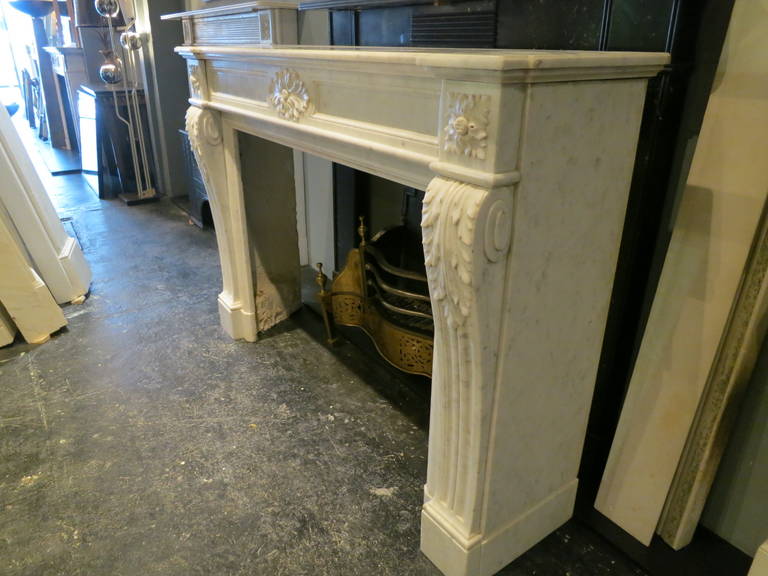 A large 19th century Louis XVI style fireplace in pale Carrara marble. Having a shaped and molded breakfront shelf, carved paterae corner blocks and carved patarae centre plaque. The jambs with acanthus leaf carvings.