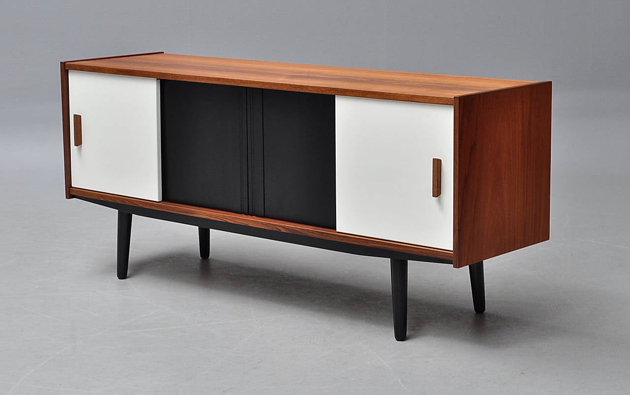 Danish furniture producer . Flat sideboard made ​​of veneered teak , front with four newly lacquered sliding doors enclosing shelves and a drawer , turned black lacquered legs . H. 66 cm . B. 150 cm. T. 42.5 cm