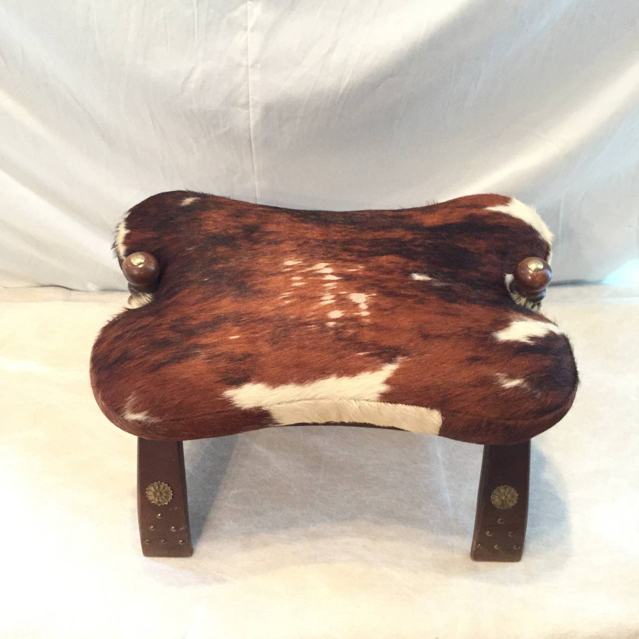 Traditional camel saddle seat upholstered in hand strewn leather and hair on hide, set atop a mahogany wood base with brass studs decoration.  Great size for a foot stool.