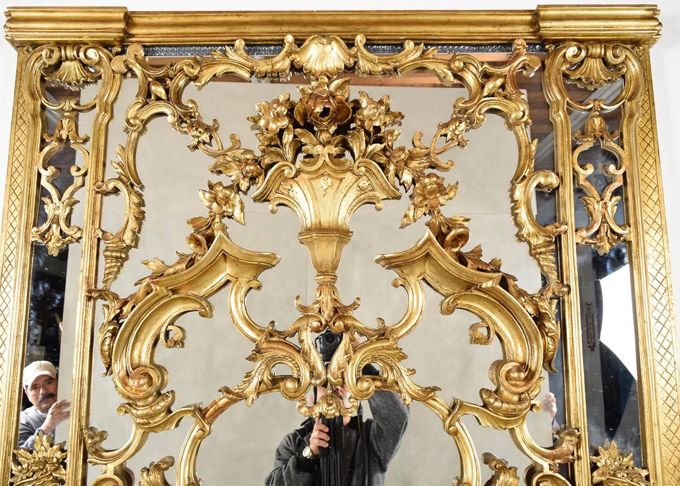 This stately 1900's Louis XVI-style large giltwood mirror will be the center of attention in any room. The solid wood frame is finished in a gold color and features heavily carved decorations of flowers, roses, scrolls, vases, leafs, and sea shells.