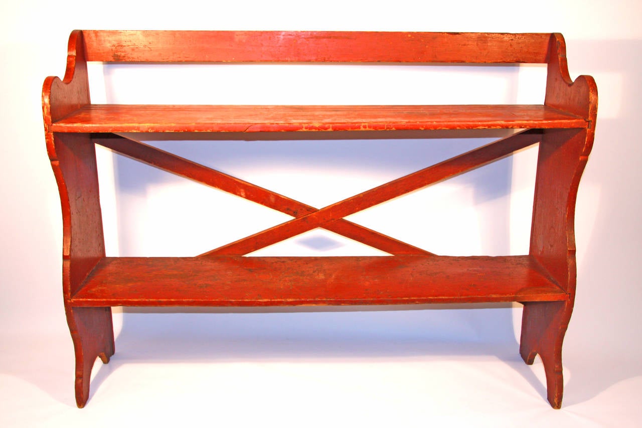 Painted pine bucket bench with two shelves, scalloped sides, and cross-braced back retaining an old red surface. Excellent condition with scalloped sides, raised feet, and cross-braced back.