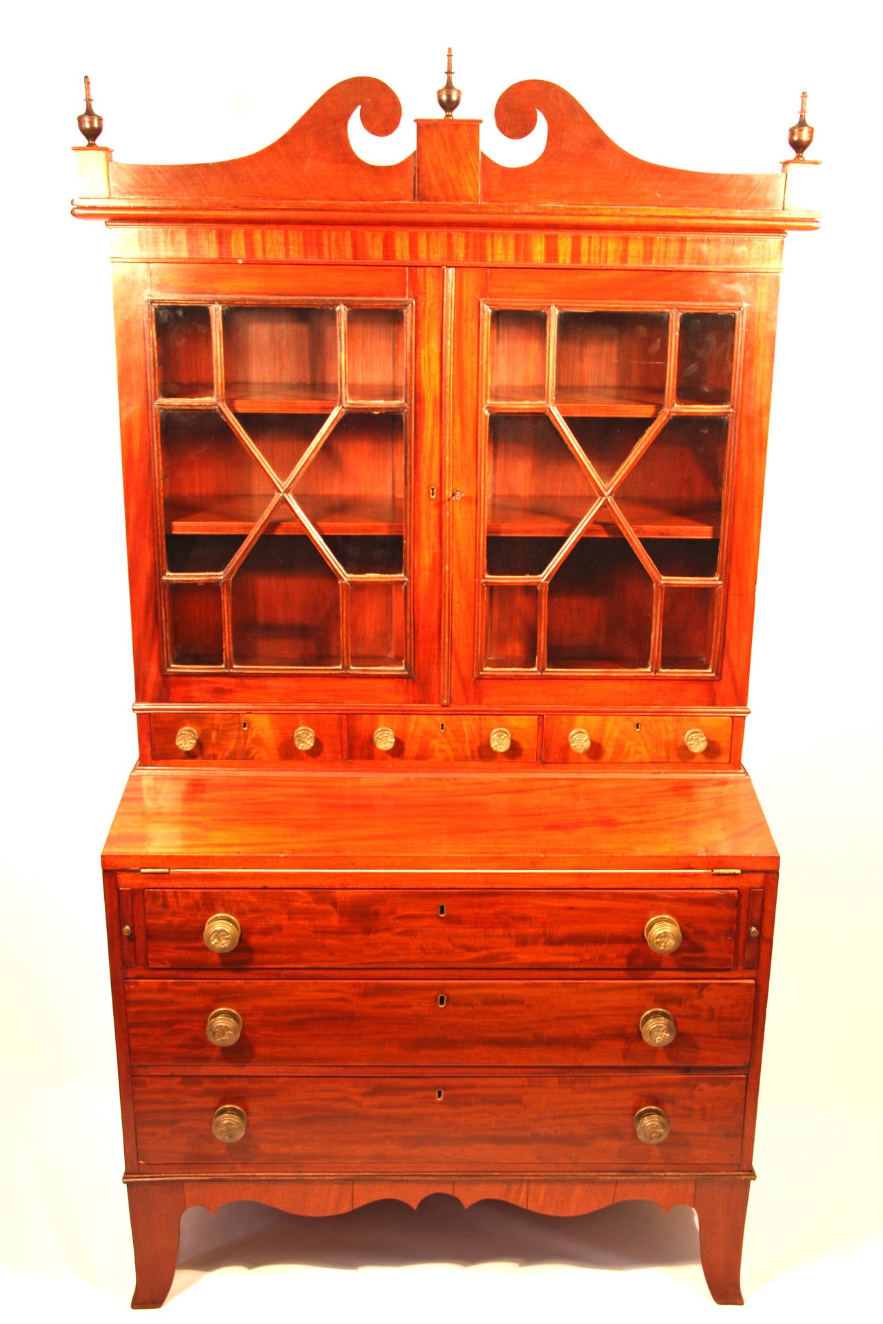 Hepplewhite mahogany secretary constructed in two parts having a scrolling arch top with plinth and finial center and ends above two multi-light doors fitted with a row of three small drawers. The base having a flip lid with two pull-out supports