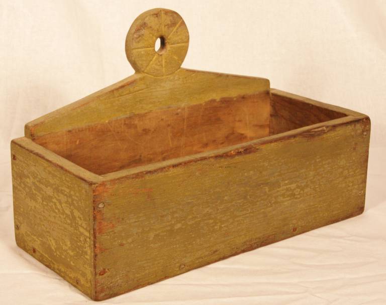 First quarter of the 19th century New England hanging wall box. The peaked backboard with pinwheel carved and pierced lollipop hanger adjoining the rectangular open box in yellow wash over original grey paint. Square nails used in construction.