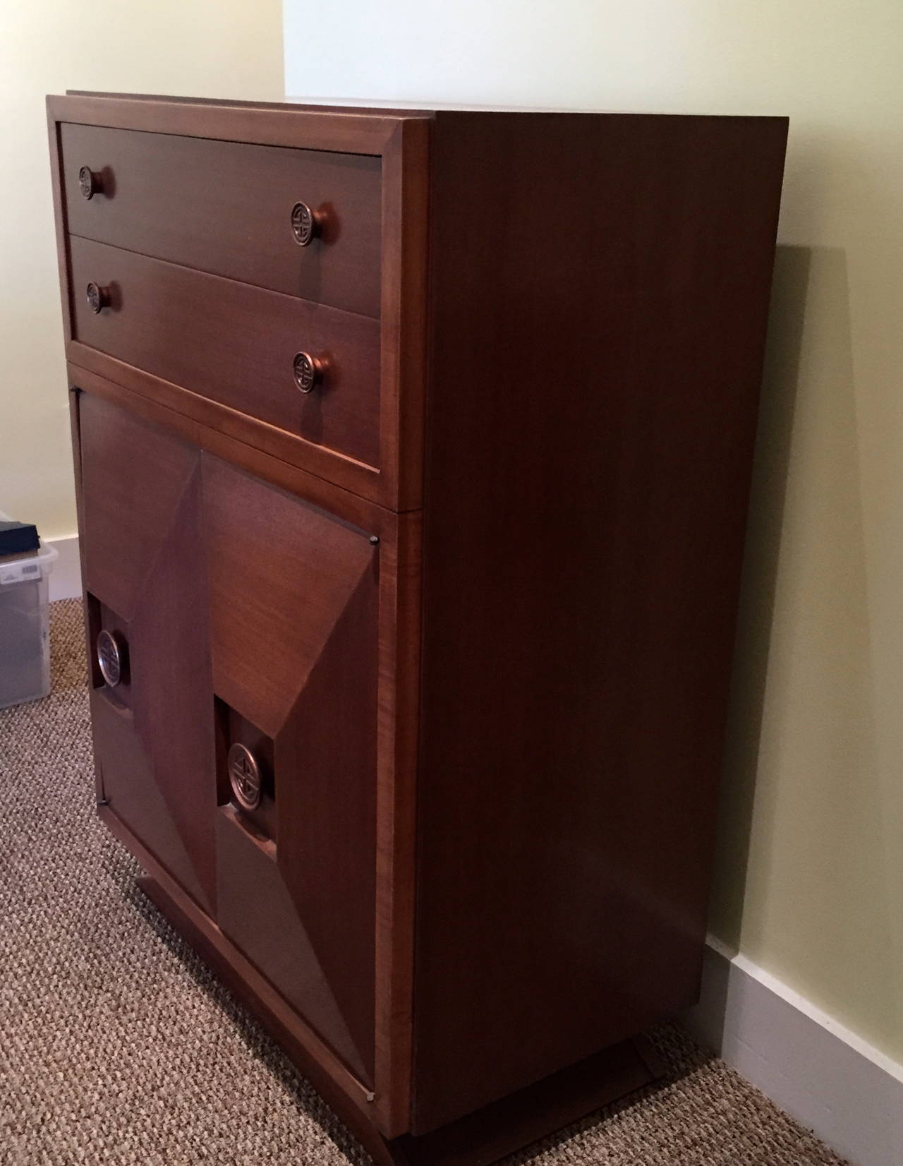 This mahogany tall chest of drawers is in the style of the great James Mont.  It has two drawers above two geometric form doors, and three drawers within the lower doors.  The hardware is Asian in design, also typical of Mont's style.

The base is