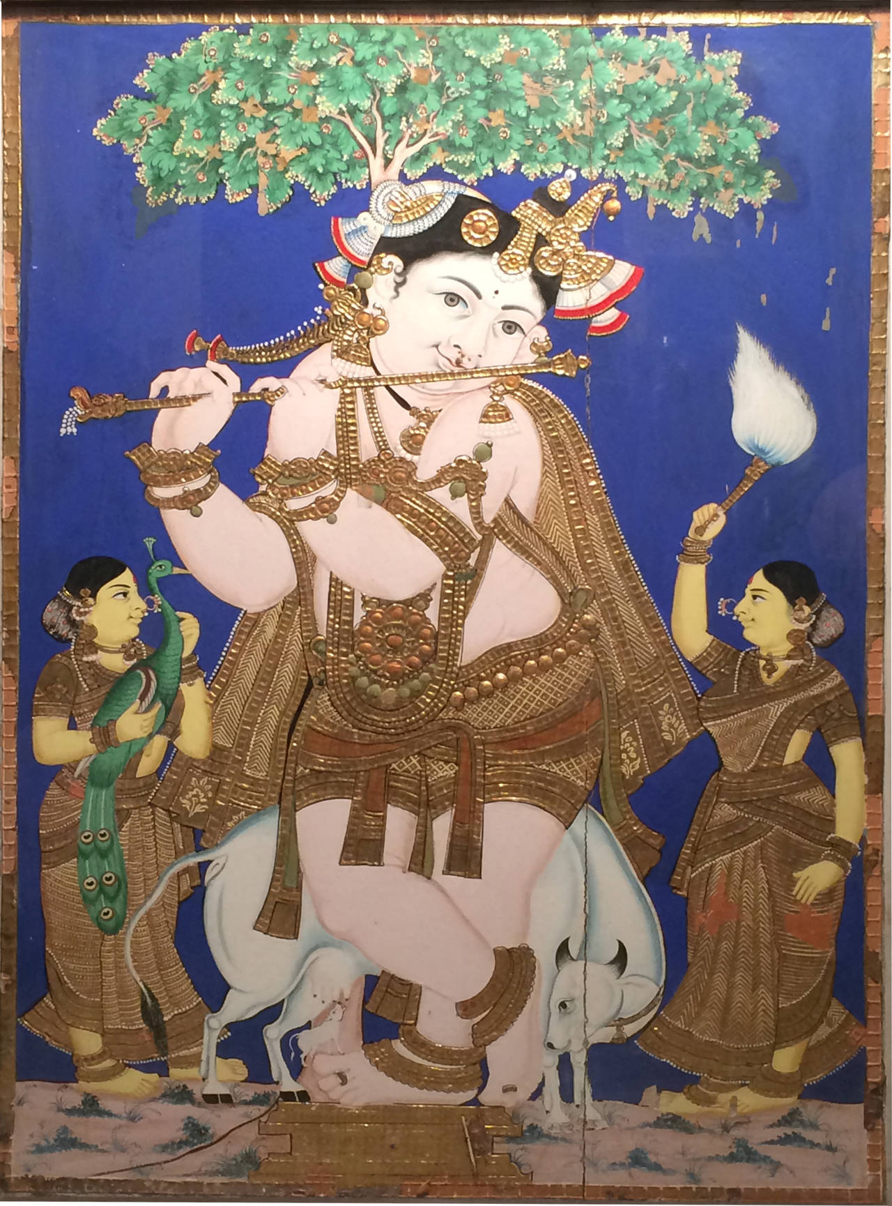 The god standing in a lively pose and holding flute to lips, a sacred cow suckling a diminutive calf below and flanked by female attendants, all figures wearing profusely gilded garments and jewelry.
South India, circa 1850.

Tanjore painting is