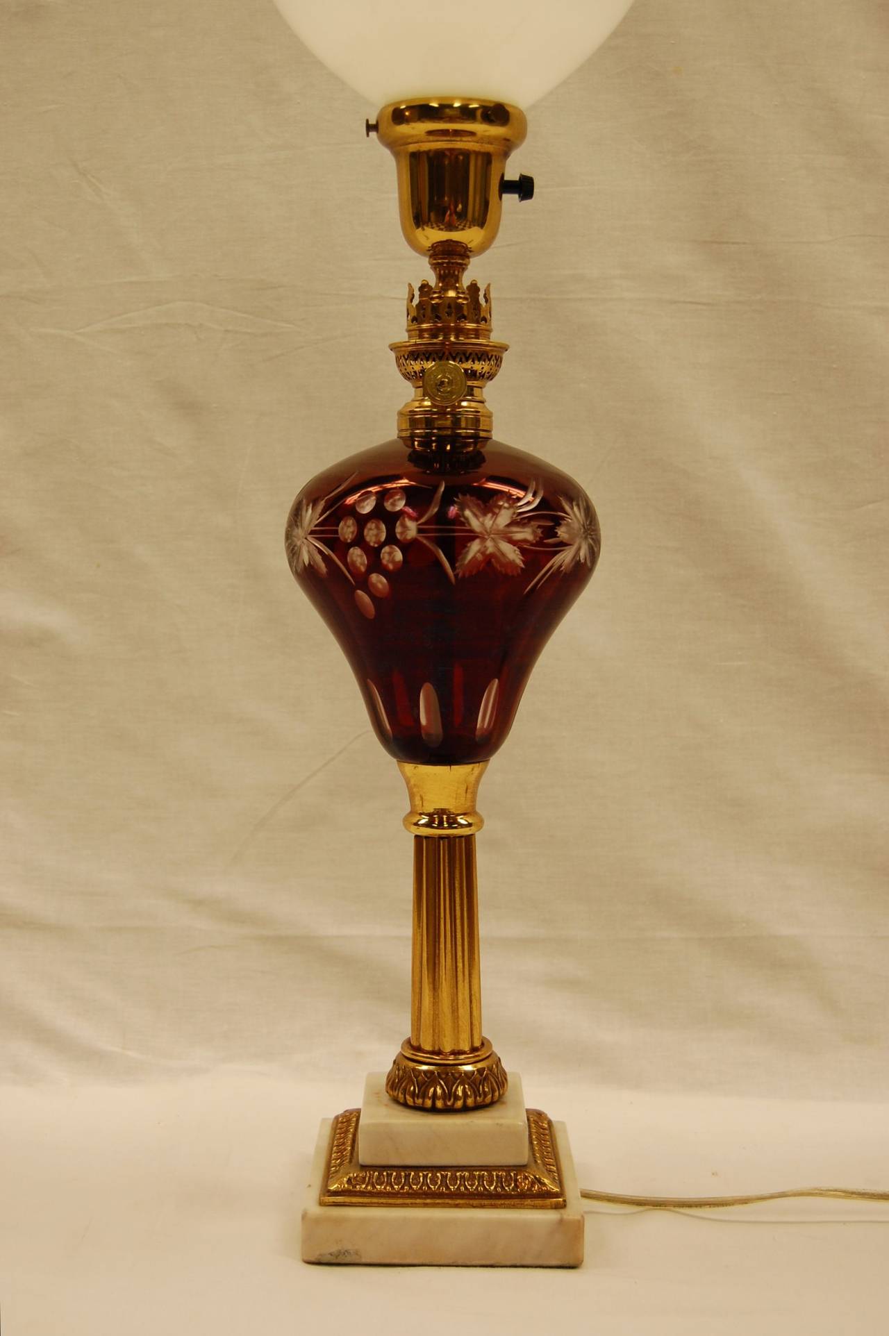 Tall ruby glass oil lamp on brass column and white marble base with brass collars. The font is cut in a grape design. Recently cleaned and rewired with a three way socket. The height to the top of the burner is 17 1/4