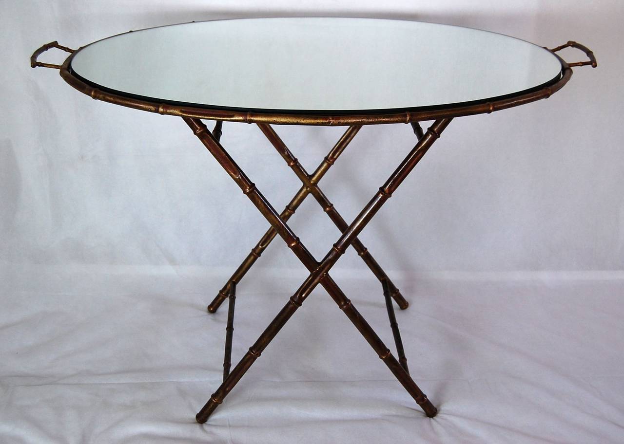 Oval top with handles on X frame base in aged gold paint imported by Yale Burge. The mirror rests (but not glued) onto a panel of plywood attached to the oval metal frame. The base can be used in the upright position, with a resting height of 28