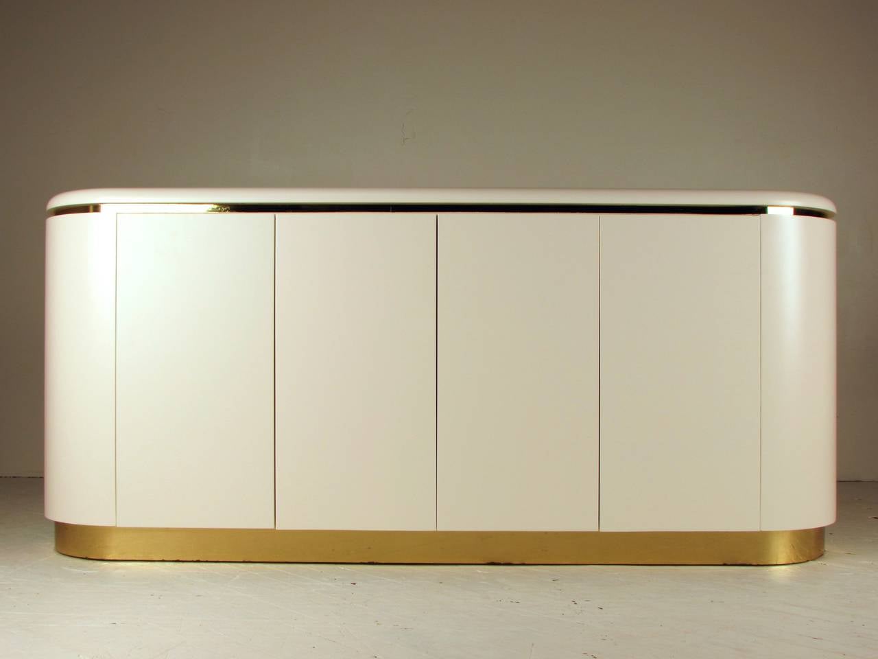 1970s glam curved buffet or credenza by Mastercraft. Impeccable quality and seriously heavy. The finish was quite dated, so we had this piece re-lacquered in Dover White, the perfect shade of off-white! 

Brass is in very good vintage condition