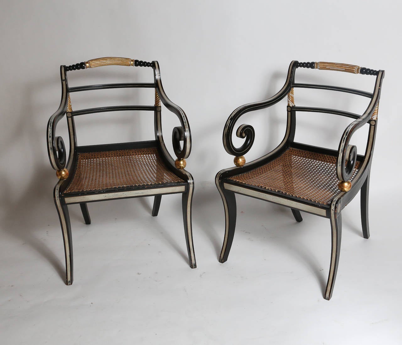 Pair of black painted English Regency armchairs with splayed legs.

Top rail and sides between back slats carved and gilt. Arms rest upon a pair of gilt balls.

Arms and frame pinstriped in grey.