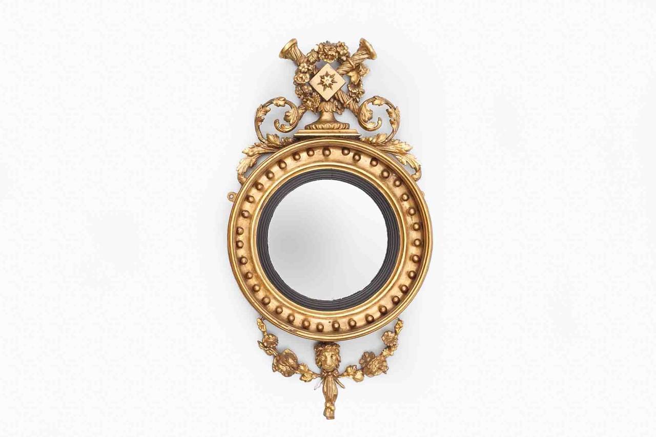 Early 19th century Regency convex mirror, the plate of circular form set within reeded slip and ball decorated surround surmounted pair intricately carved pair of trumpets and star embelem with foliate garland of flowerheads flanked with scrolling