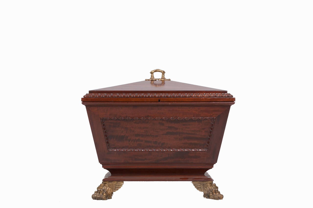 Early 19th Century mahogany wine cooler of sarcophagus form, the raised hinged top above the body with recessed panels with carved legs on a platform base with winged lion paw feet of gilt metal.