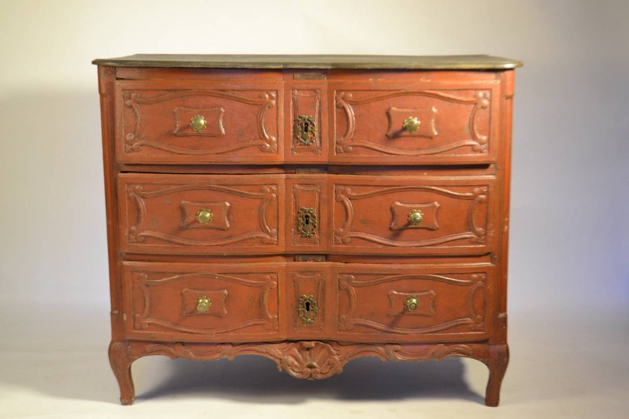 A rare Louis XIV/Regence commode newly painted in ox blood red, with three convex and concave scrolled molded panels with beveled molding and central brass pulls on the drawers, with a central pierced bow and floret motif on the key plates. On the