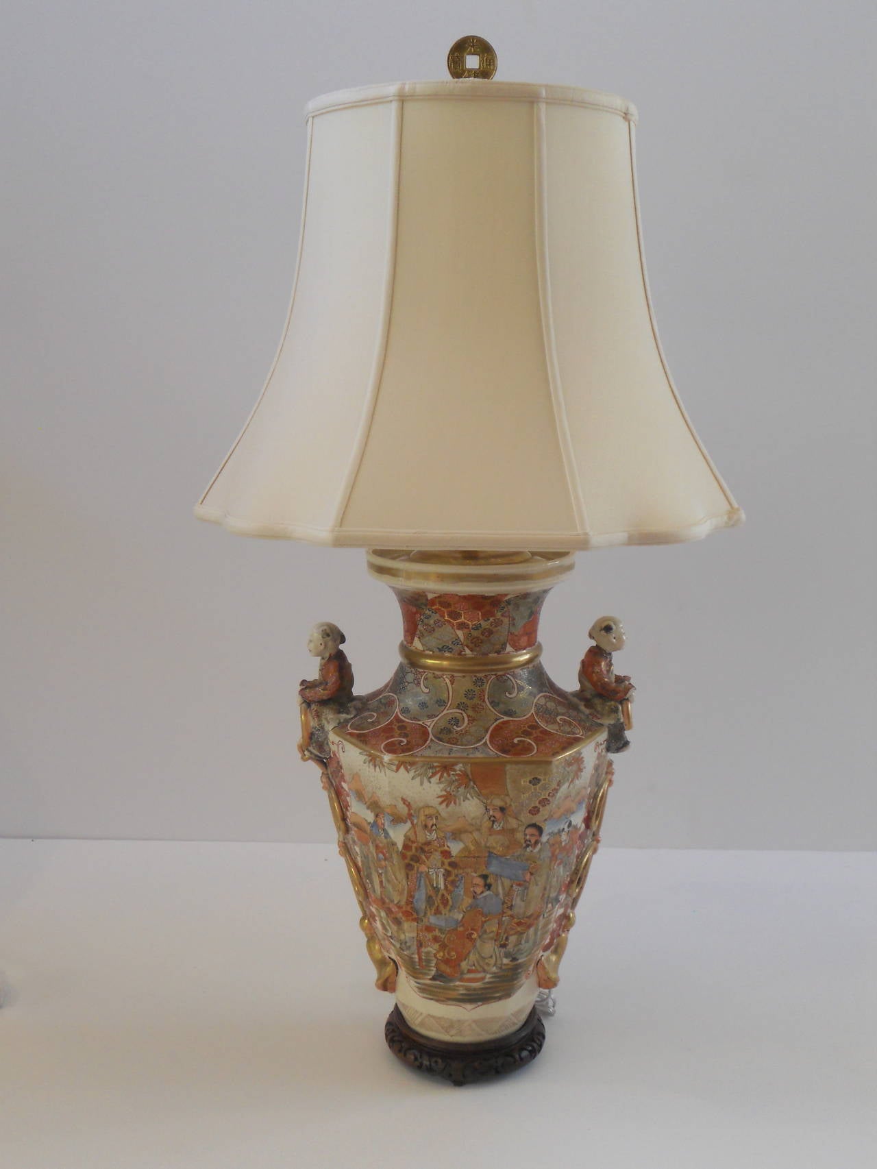 Large Satsuma vase as a lamp with a pair of figures resting atop the urn form vase holding gold interlocked tassels. The piece is hand-painted with highly detailed decoration and figures in soft shades of orange, pale blue, gold and deep, rich red.