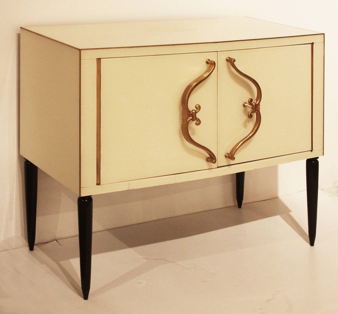 Sophisticated Mid-Century Modern French cabinet in glass over bone colored, painted wood with handsome bronze detailing and gilt bronze door hardware and elevated on ebonized legs. A closely related cabinet is pictured on the cover of