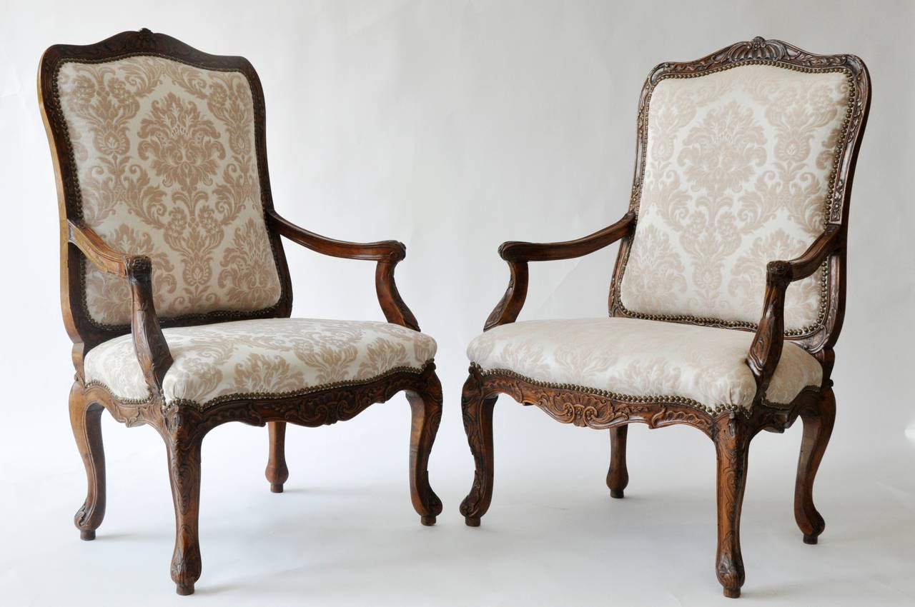 Beautiful pair of 18th century Louis XV walnut armchairs.

Detailed hand carving to top, front apron and both arms and legs.

Chairs upholstered in neutral ivory and taupe cotton damask. Antique brass nailhead trim around seat back and bottom.