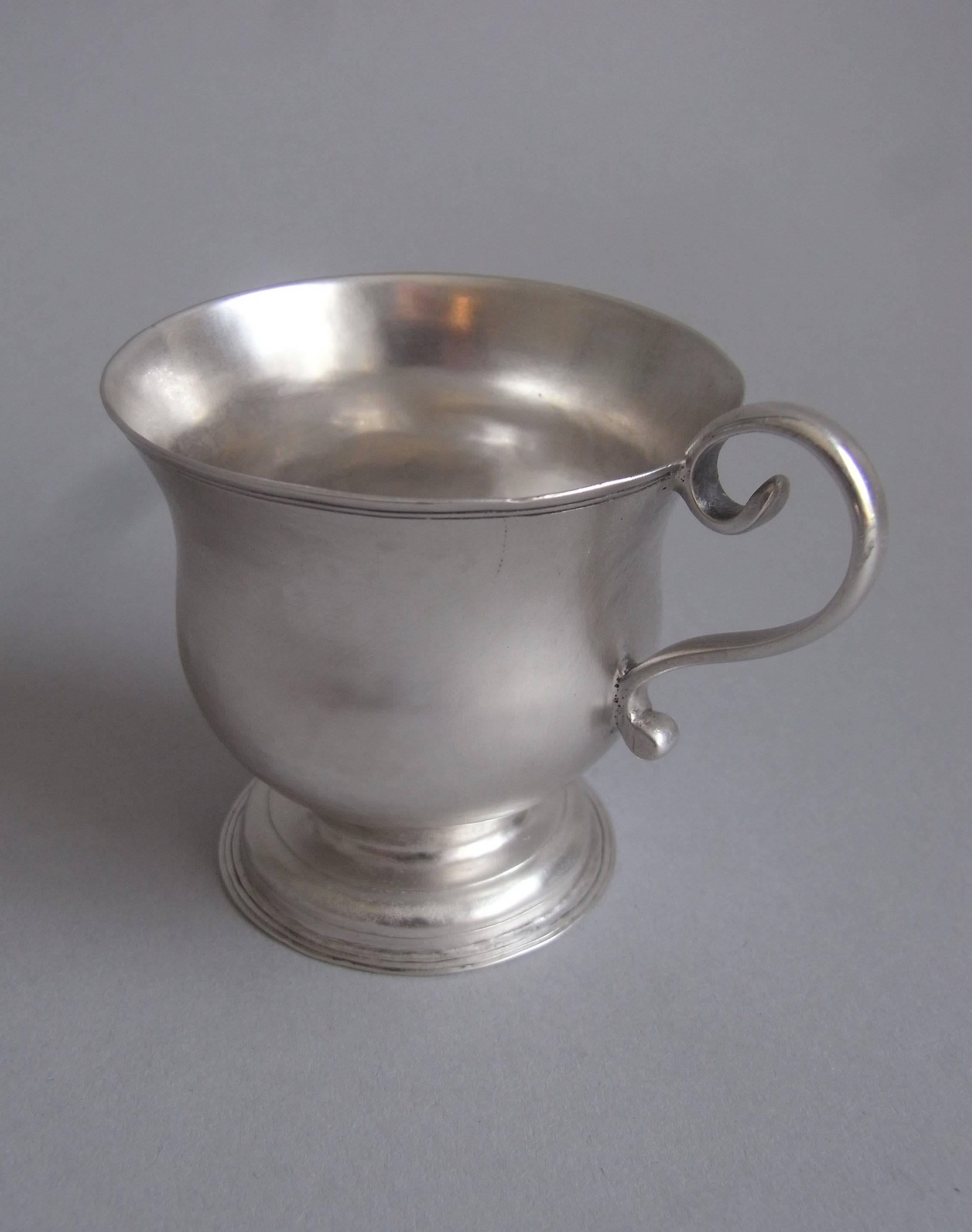 The cup stands on a stepped circular foot decorated with reeding. The bell shaped main body has an everted rim and is plain in design. This example has an attractive scroll handle and is engraved in the foot with a set of contemporary scratch