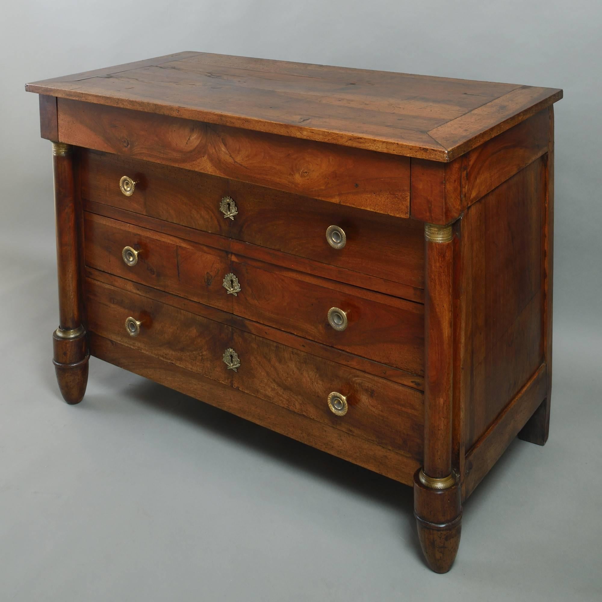 An early 19th century Empire Period walnut commode, the cleated top above a hidden frieze drawer and three long drawers with book-matched veneers and finely chased handles and lock escutcheons, between two turned column supports, all standing upon