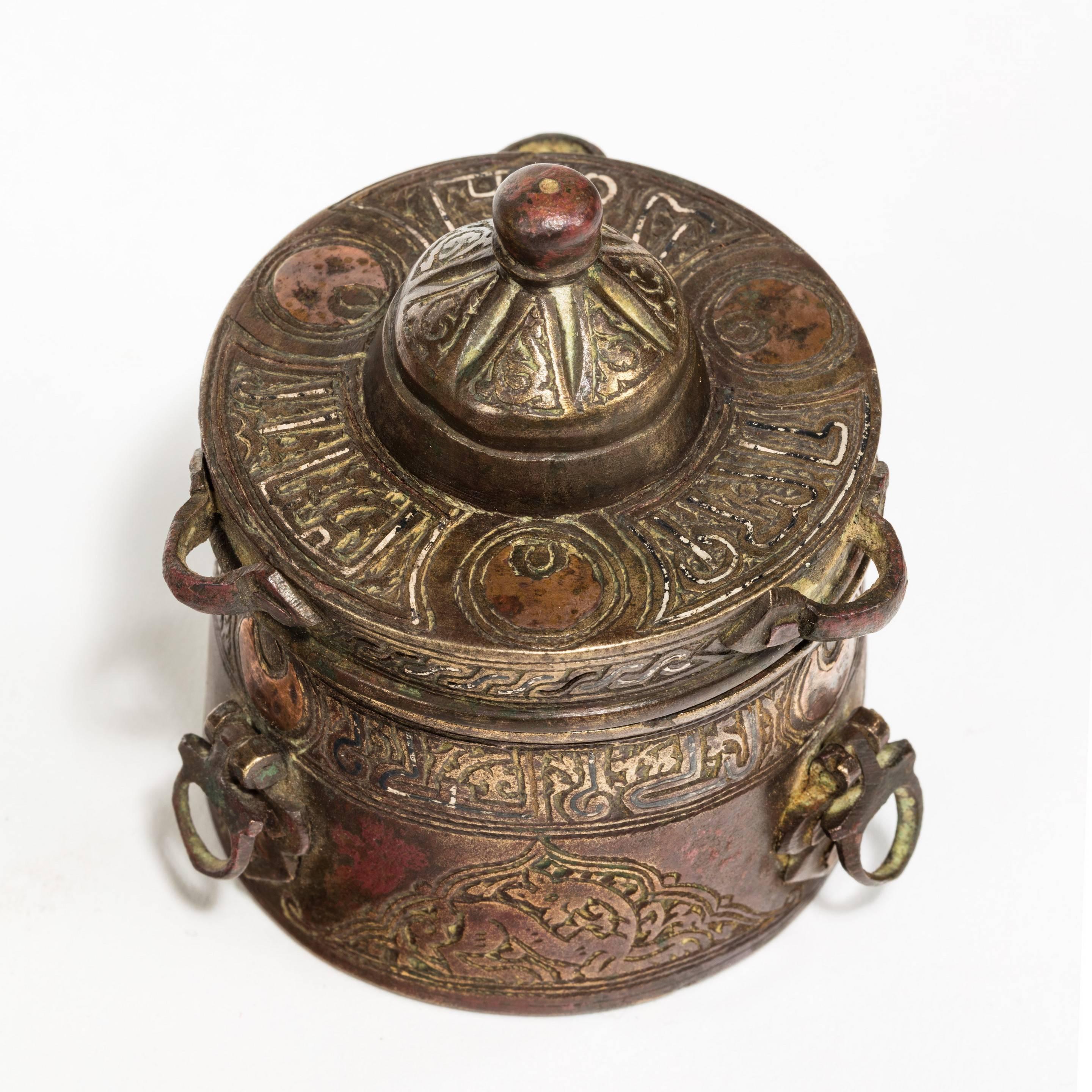A Khorasan bronze inkwell, of typical form with three small handles and a lobed cover (hinge missing), inlaid in copper and silver with bands of calligraphy and figural roundels, 12th century.