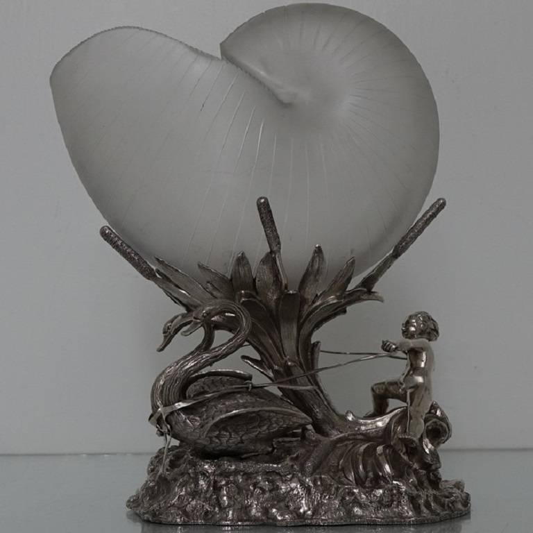 A stunning silver plated centrepiece with a glorious contemporary nautilus themed glass shell. The body is decorated with a cherub riding a swan drawn chariot. The centre glass dish is held aloft with a beautiful foliage trunk. 

Length: 6