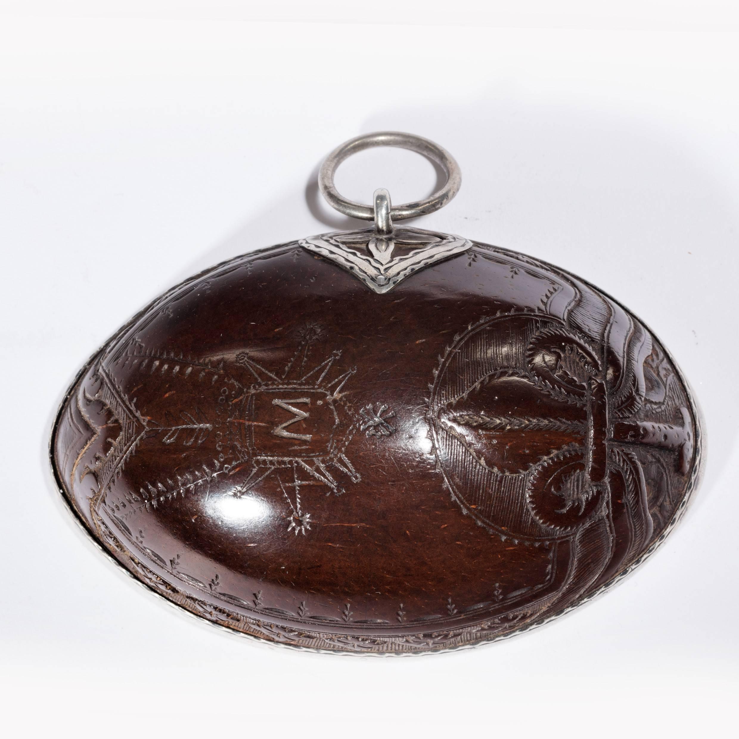 A Portuguese colonial coconut shell scoop made from half a coconut with silver mounts and attachment ring, incised with a fleur de lys and the initial ‘M’ in a shield, circa 1800.