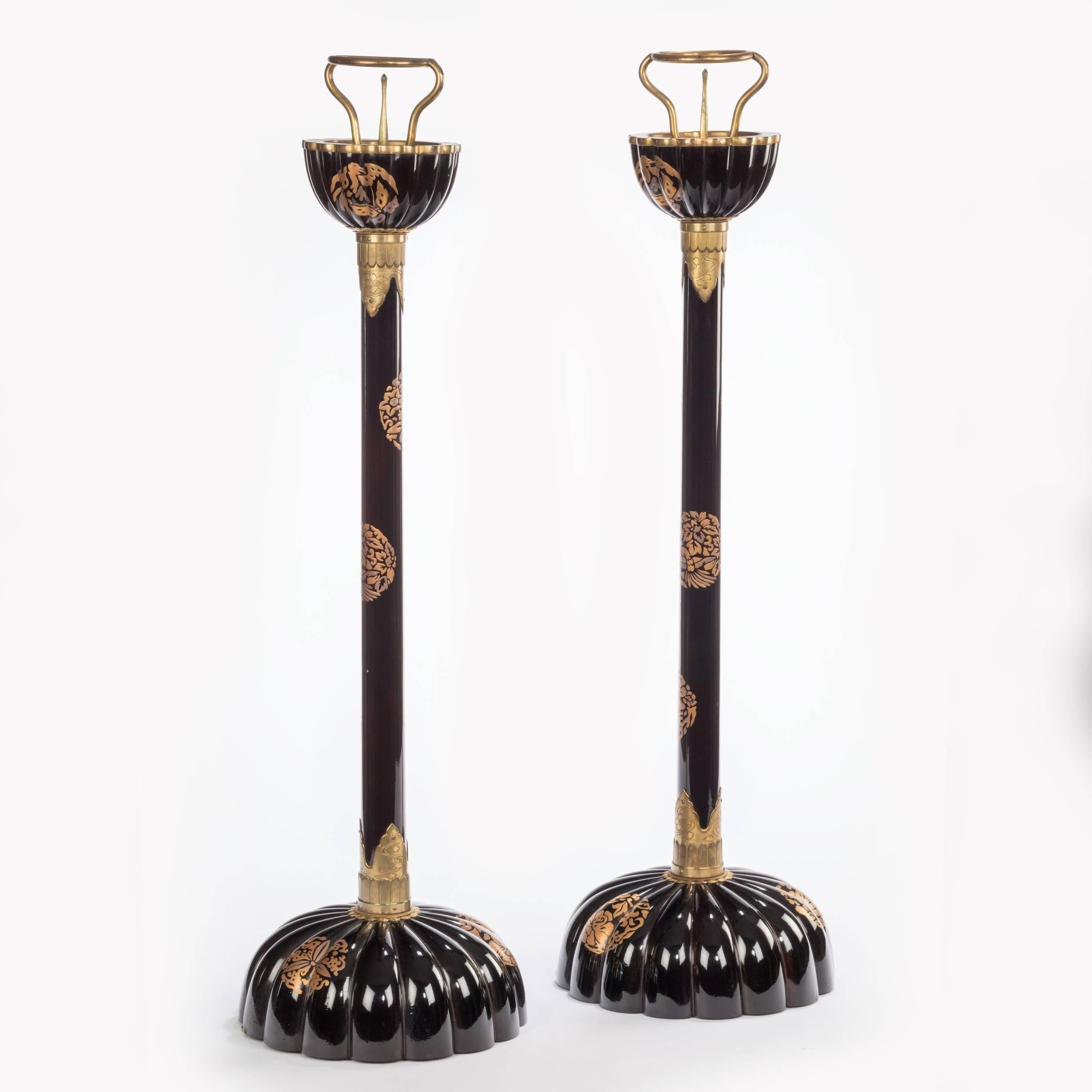 Each in with a cylindrical column rising from a stylized chrysanthemum base with a similar finial crowned by a gilt bracket and pricket for the candle, with an incised gilt metal collar and decorated with gilt mon on a black ground, with a large