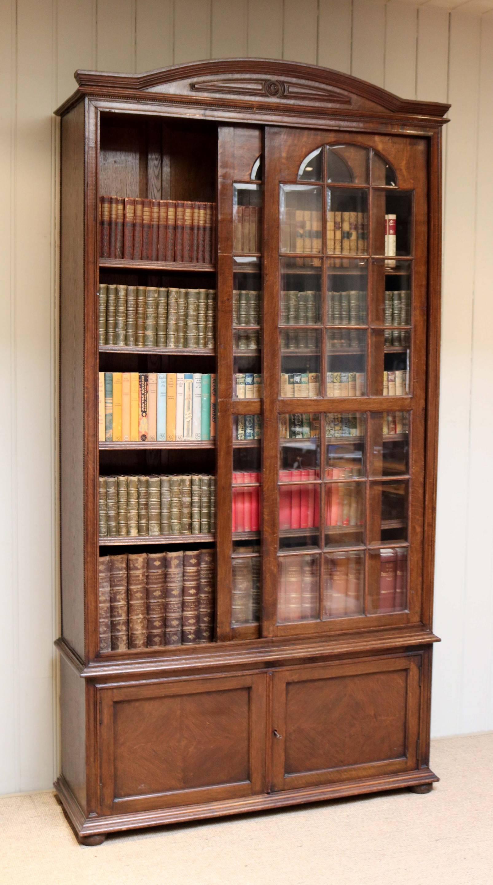 Solid oak glazed bookcase, with an arched top, having glazed bevel edge sliding doors, give access to four fully adjustable internal shelves. Sitting above a two door cupboard, supported on bun feet. 
Shelf width 22 cms.