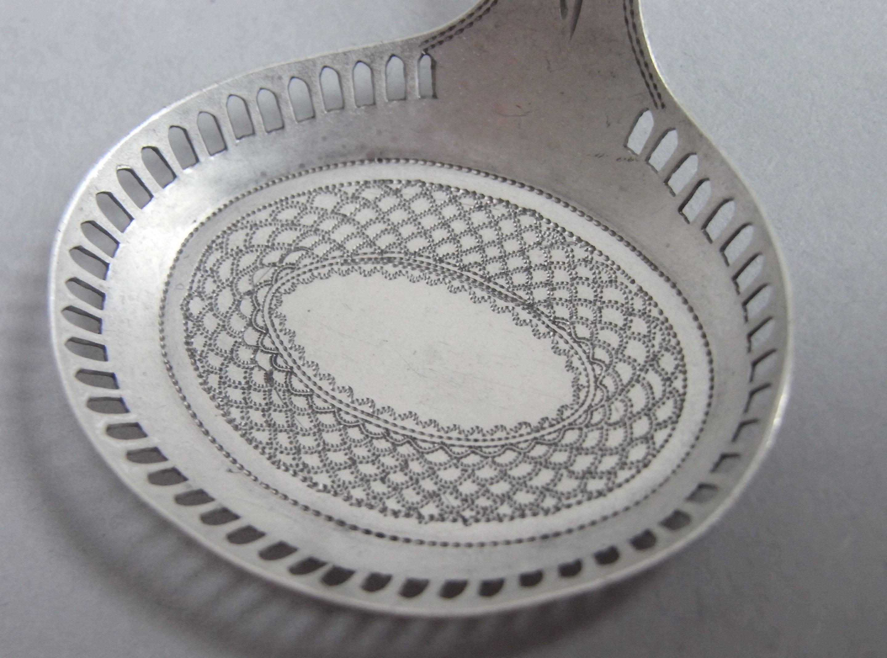 The caddy spoon is modelled as a frying pan with oval bowl, displaying raised sides with a pierced gallery. The bowl is engraved with a wide band of prick dot concentric designs around a vacant oval. The curved handle is beautifully engraved with