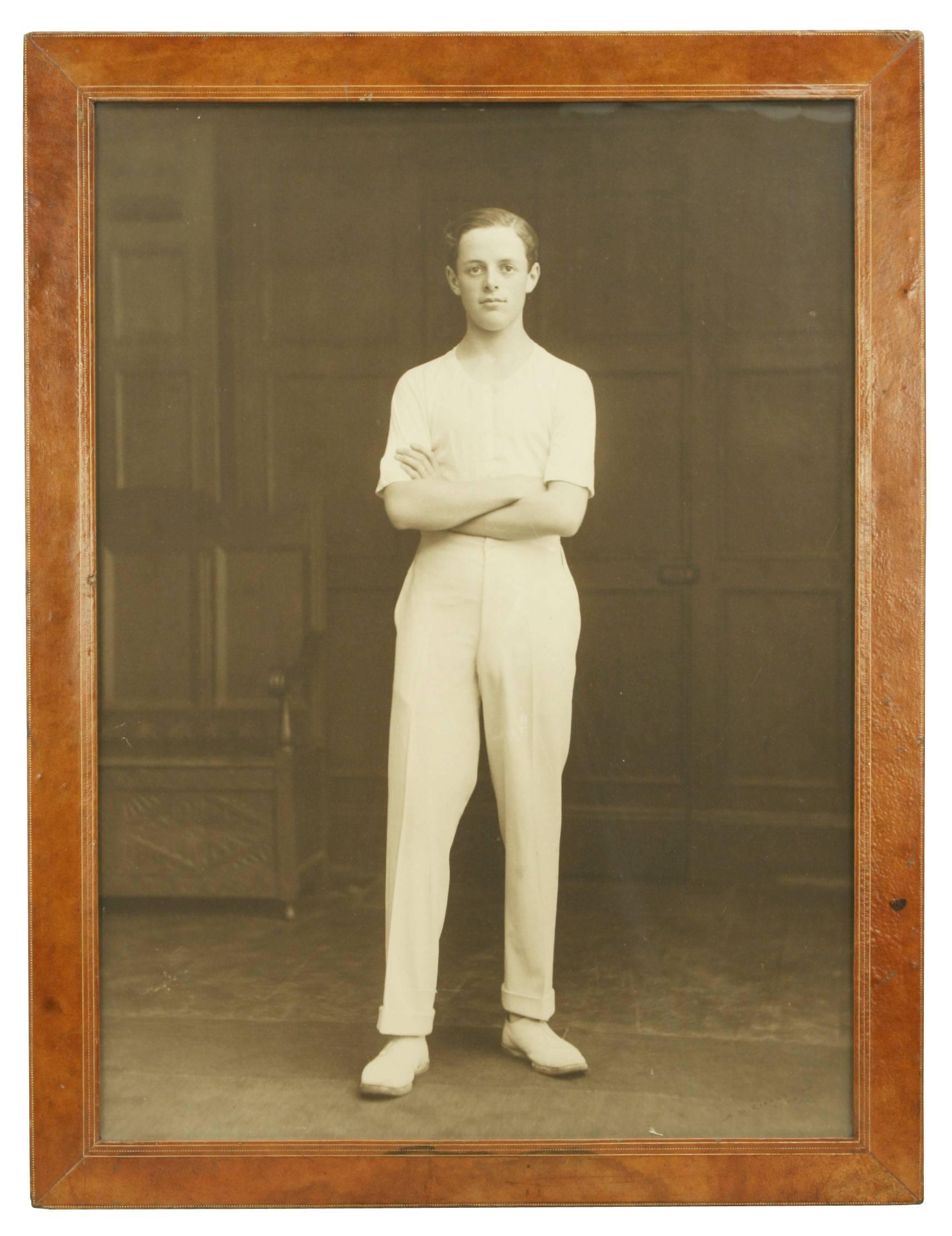 Large studio photograph in a wonderful tan leather frame. The studio photograph, full length, is of a standing young man/schoolboy, his arms folded, wearing pale short sleeved sports vest with white trousers and shoes, subject unknown. He is stood