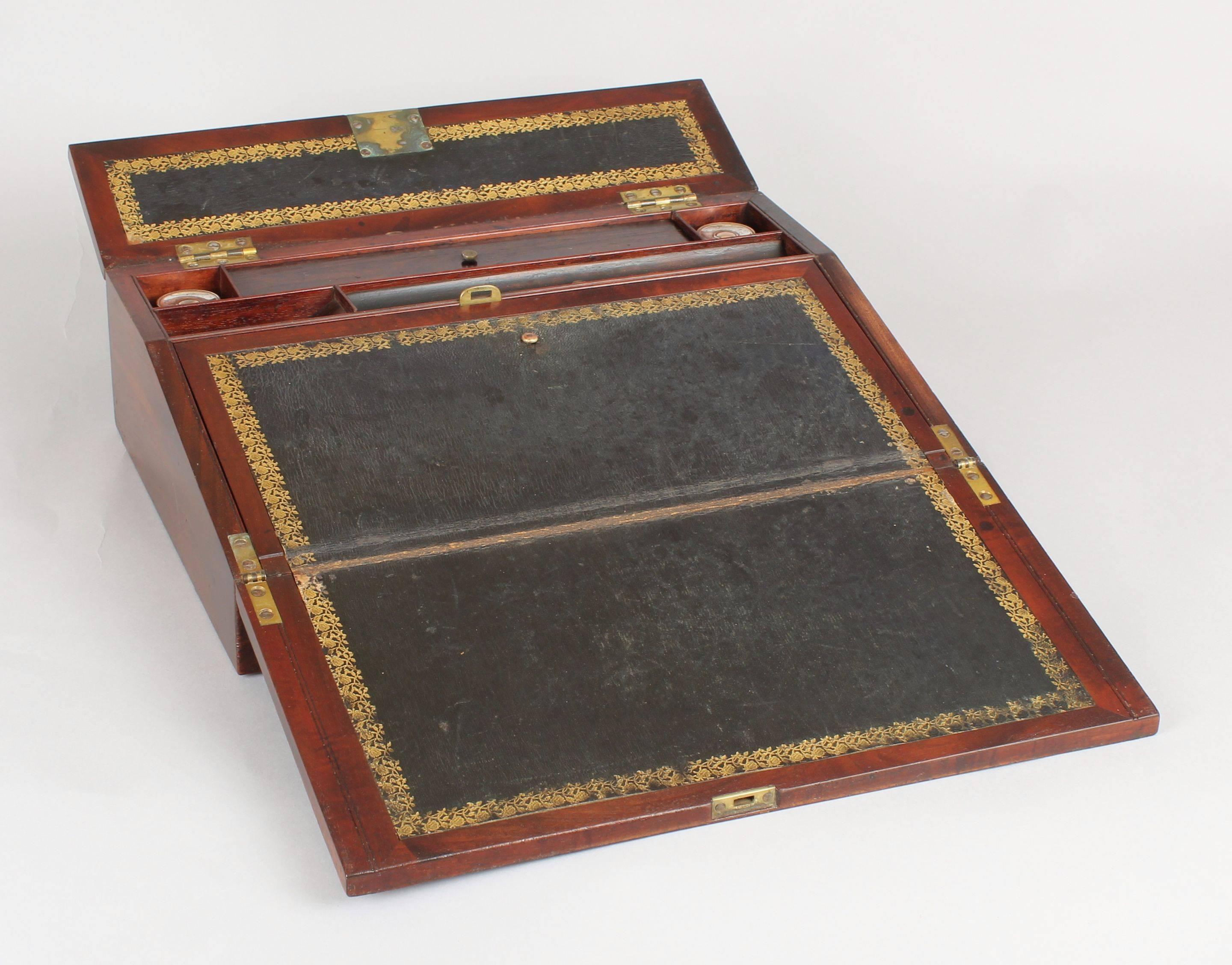 William IV period mahogany lap-desk; the hinged lid and writing-slope inlaid with cut-brass borders and a flush-fitting handle; the desk with original gold-tooled green leather flaps enclosing two pen-boxes, a nib-slop and a pair of inkwells with