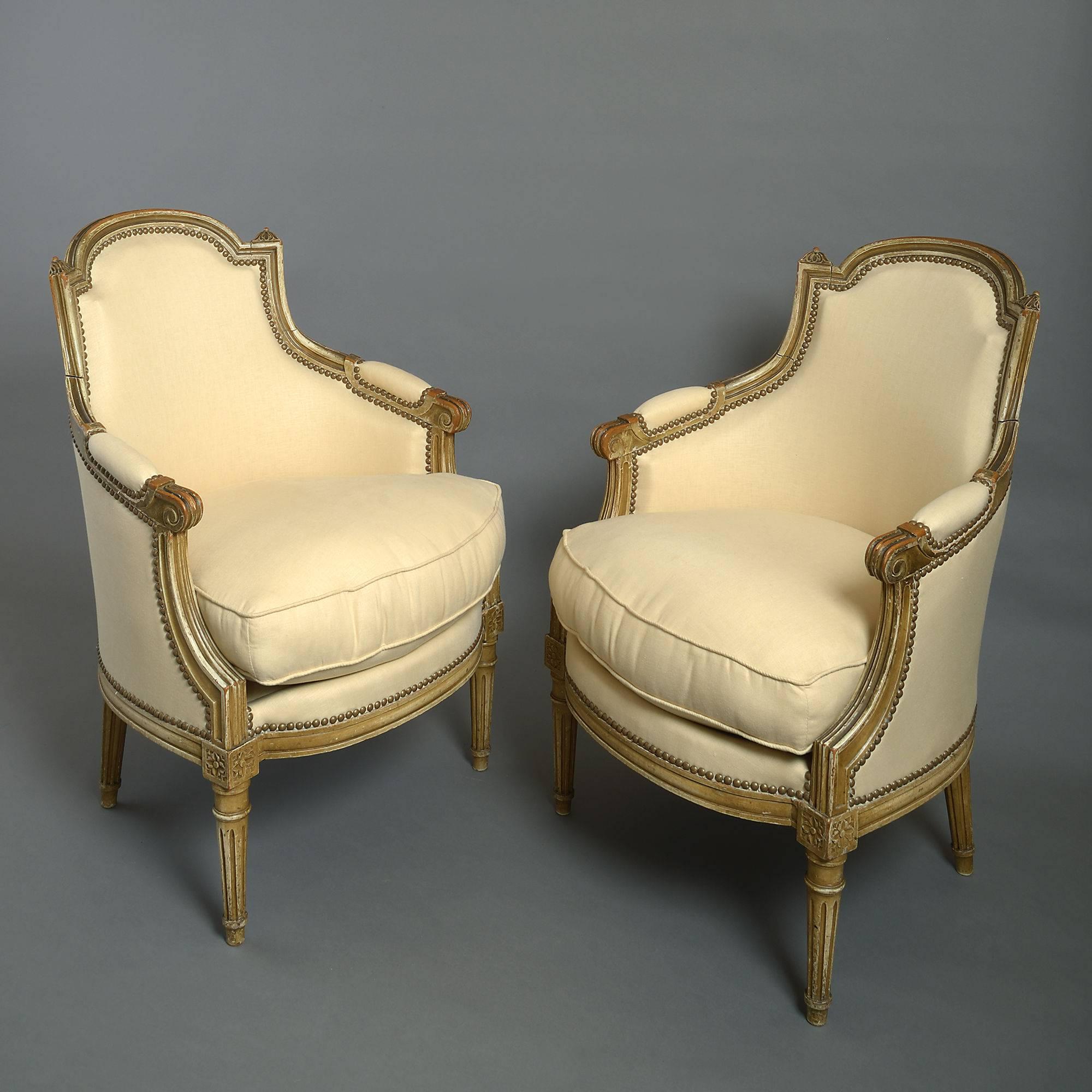 A late 19th century pair of painted bergère armchairs in the Louis XVI manner, the arched backs with finials, scrolling arms and all raised upon turned tapering legs. 

Upholstered in a cream linen with patinated brass studs.