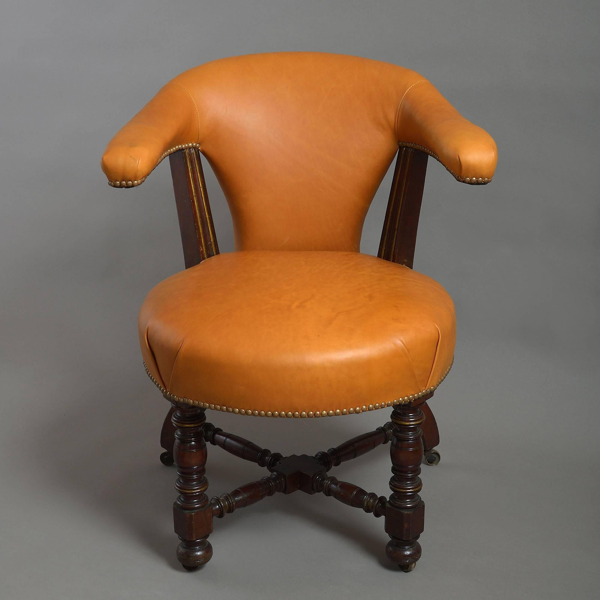A late 19th century Victorian period mahogany and parcel gilded reading armchair, the back, arms and seat all upholstered in tan leather with patinated brass studding, the whole raised upon turned legs conjoined with an X-form stretcher and
