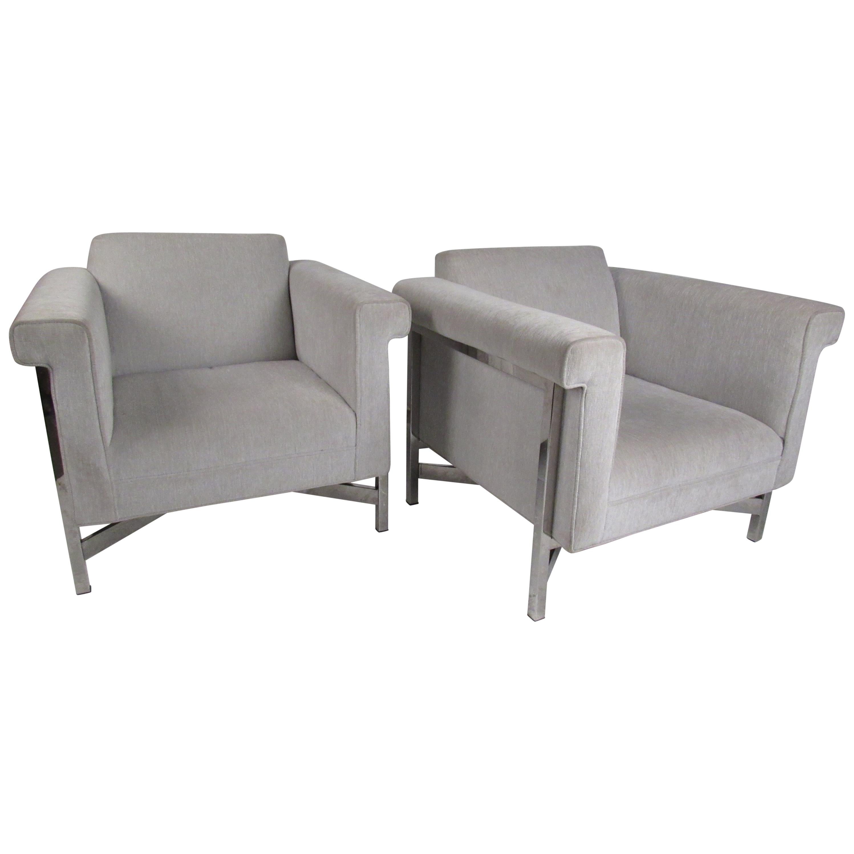 Este impresionante par de sillones de salón modernos y contemporáneos tienen armazón cromado y un tapizado afelpado de color gris claro. Un diseño elegante con reposabrazos alados, asientos bajos y camillas en forma de 