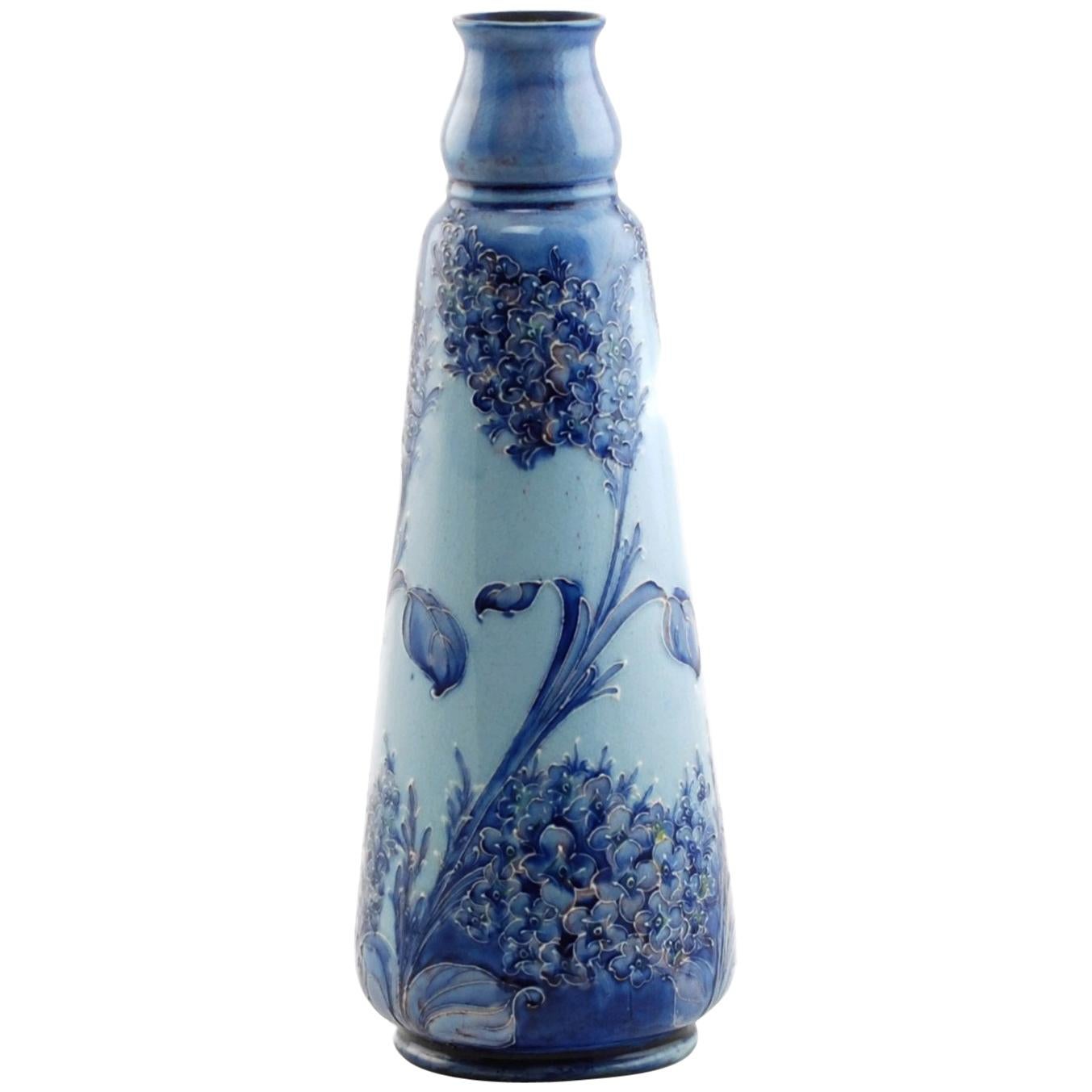 This English Art Nouveau hand decorated Florian Ware vase was designed by William Moorcroft for James Macintyre of Burslem England. The piece has a tapered form and features tube lined decoration executed in the charming Lilac pattern. The dark blue