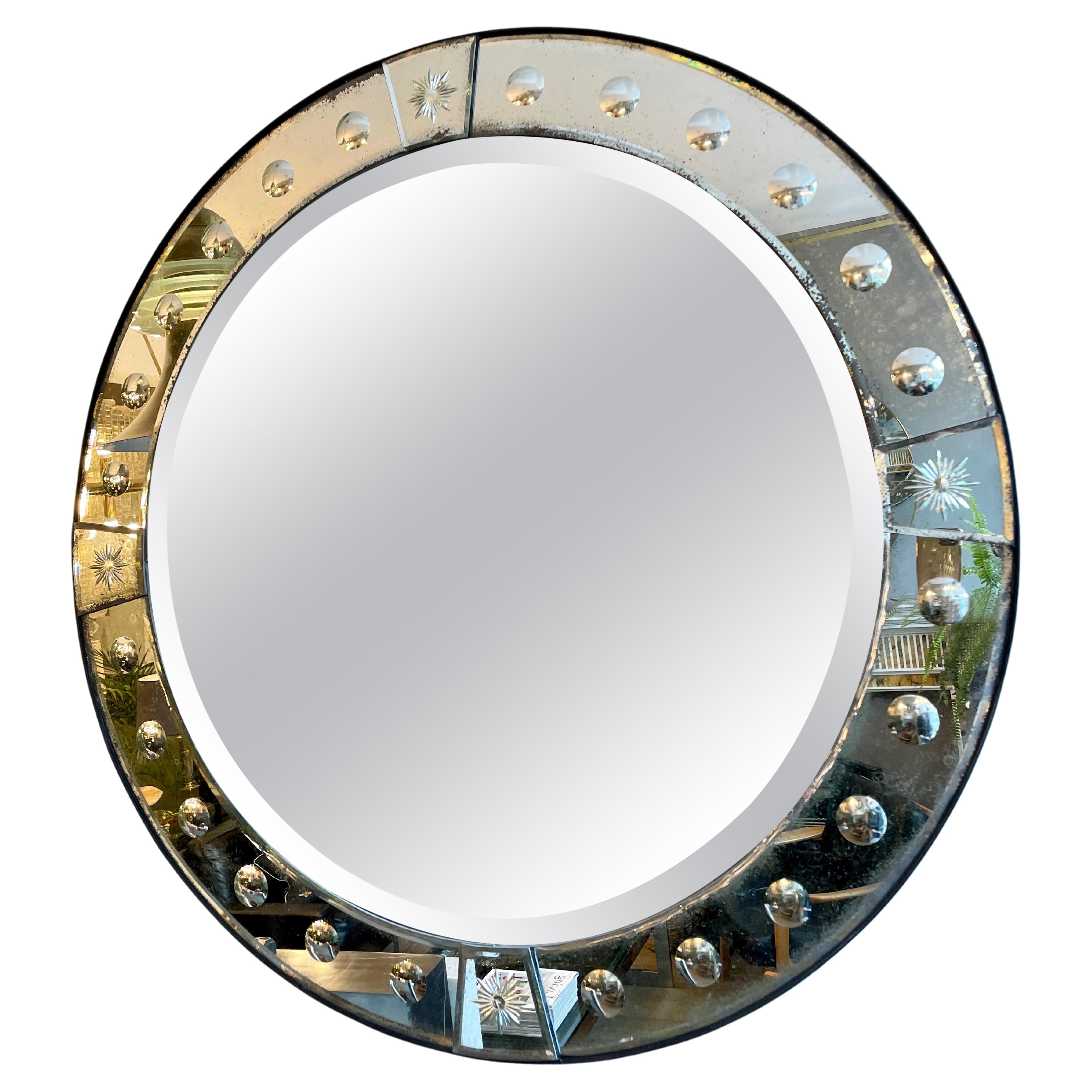 Un miroir circulaire avec une bordure de miroir à panneaux en tôle légèrement vieillie, monté sur du bois qui a un bord en bande d'acier ébénisé. Décorée de verres circulaires gravés et biseautés.