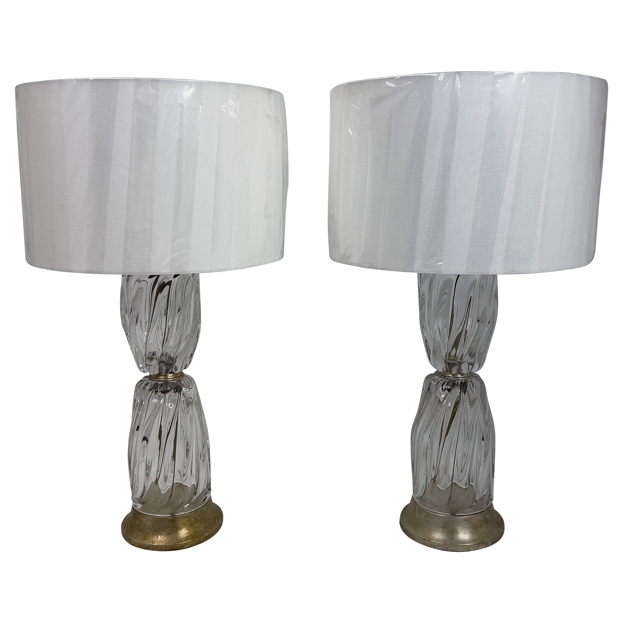 Monumental Pair of Italian Murano Clear Glass Lamps. The glass has a twisted pattern and the metal base and cap have a modeled silver leaf finish. These lamps are topped with round linen shades.