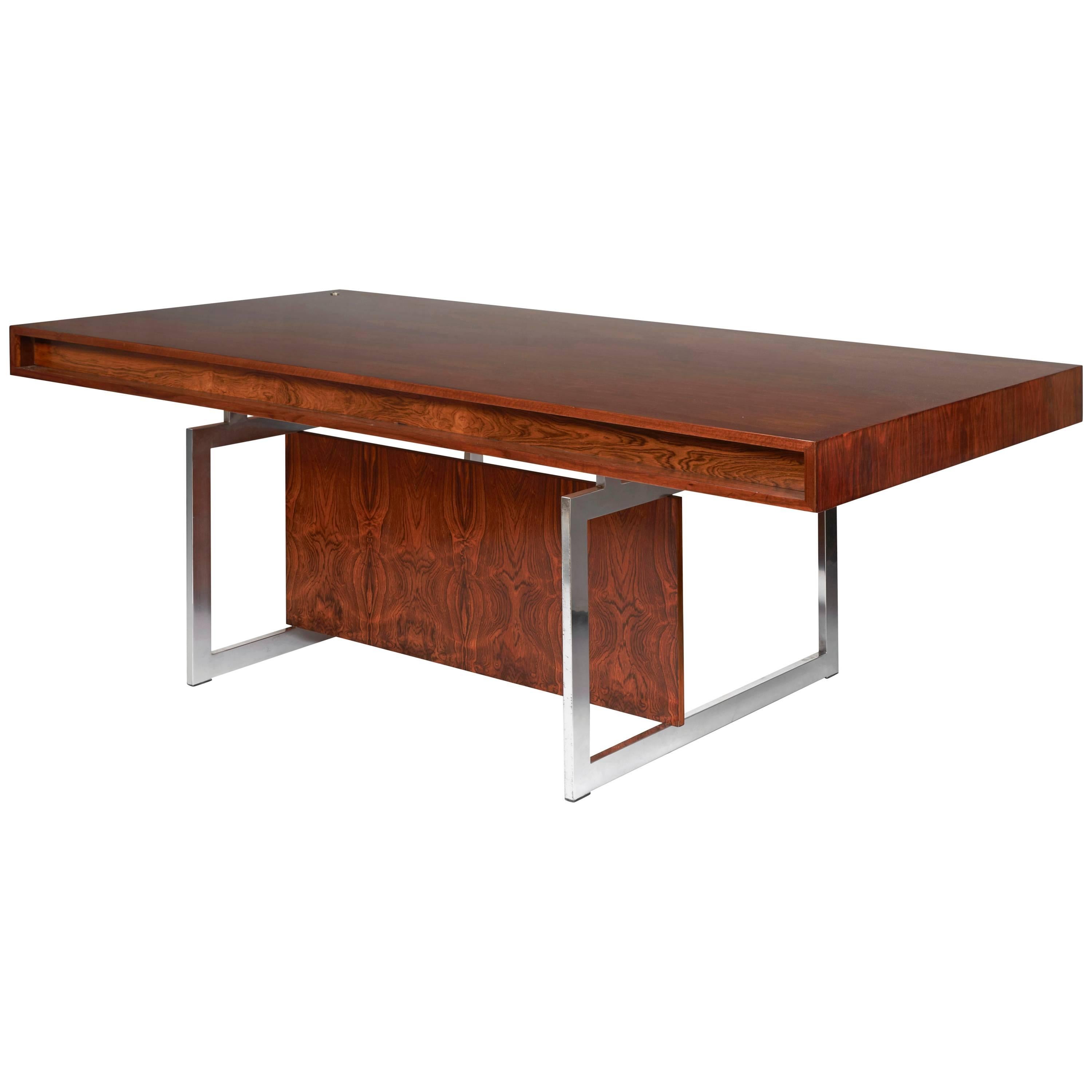 Comprising a free-standing rosewood rectangular desk with chromium-plated steel frame, the legs joined under the top by a rosewood panel, the top with four integrated drawers
with a matching rosewood cabinet on wheels. 
Manufactured by E. Pedersen