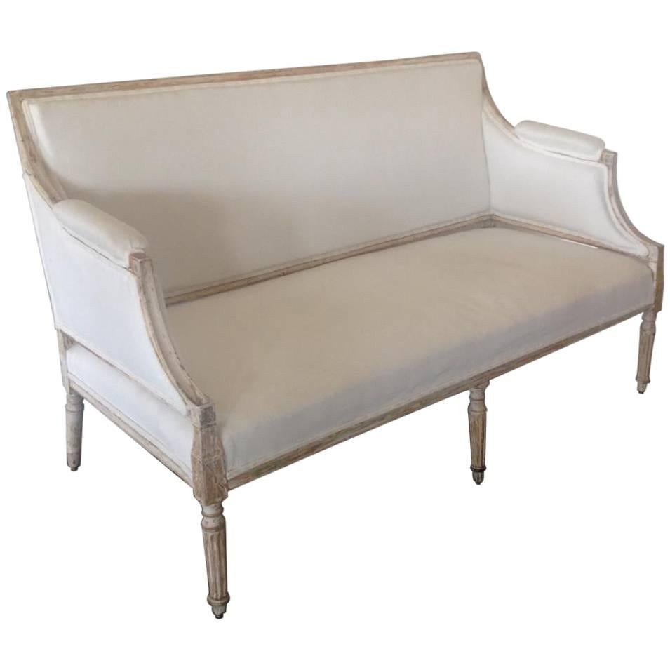 A Swedish banquette sofa hand-scraped to original chalky white Gustavian paint with traces of old gilt from the 19th century. The upholstery fabric is a crisp white Belgian linen. A very beautiful and sturdy piece. The arm height is 28".

The