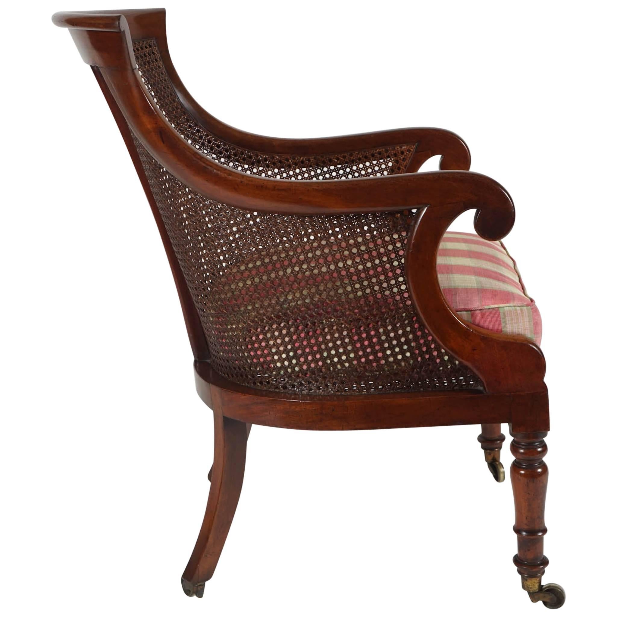 An elegant English Regency period, circa 1830, armchair, library chair, or bergere of solid mahogany construction having shaped crest rail and scroll arms with caned back, sides, and seat with loose squab cushion on turned front legs and splayed