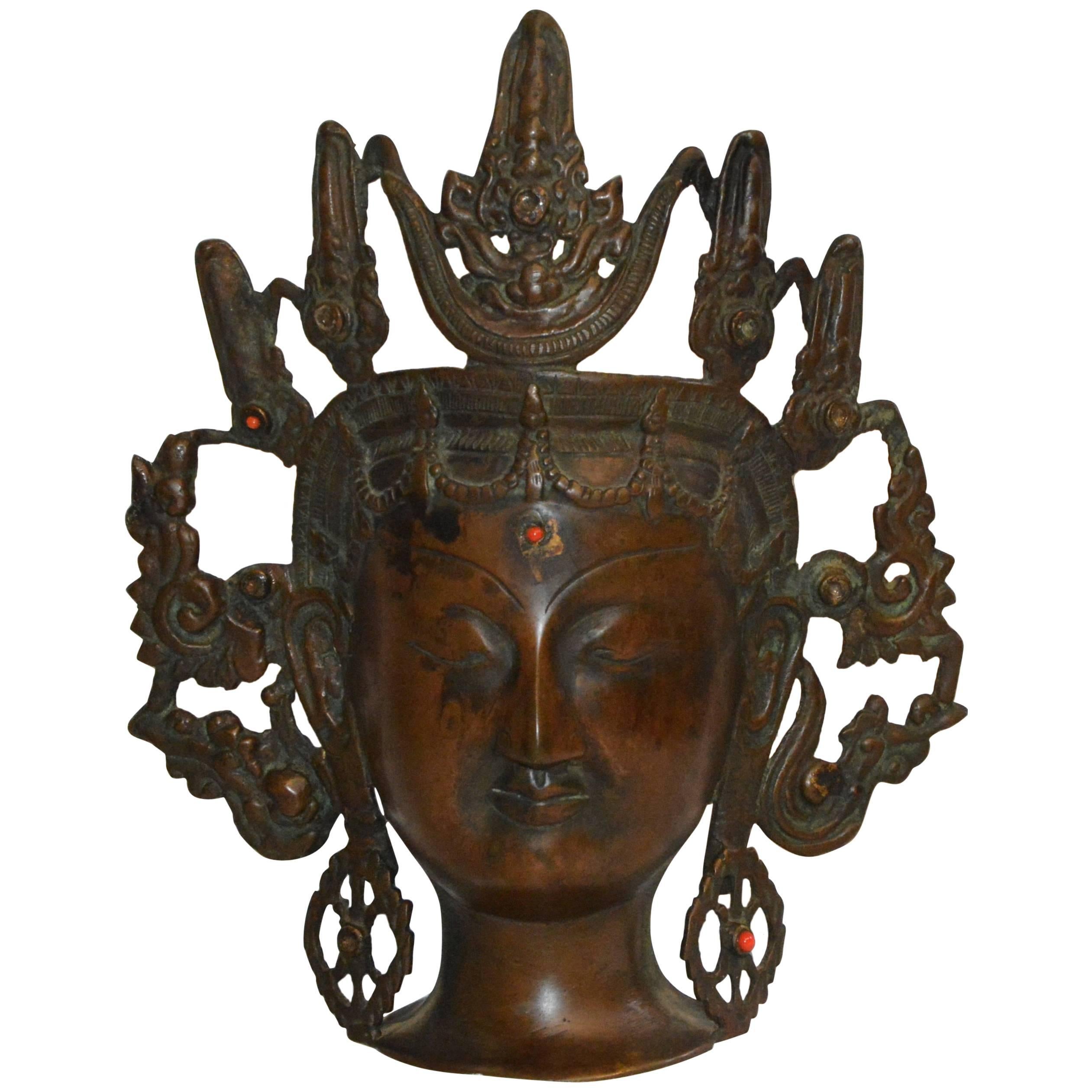 This is a Thai bronze head figurine with an ornate open work headdress. She has elongated ears with decorative earrings and a scallop border around her face. Red beads decorate her forehead and her left earring. It is marked India on the back.