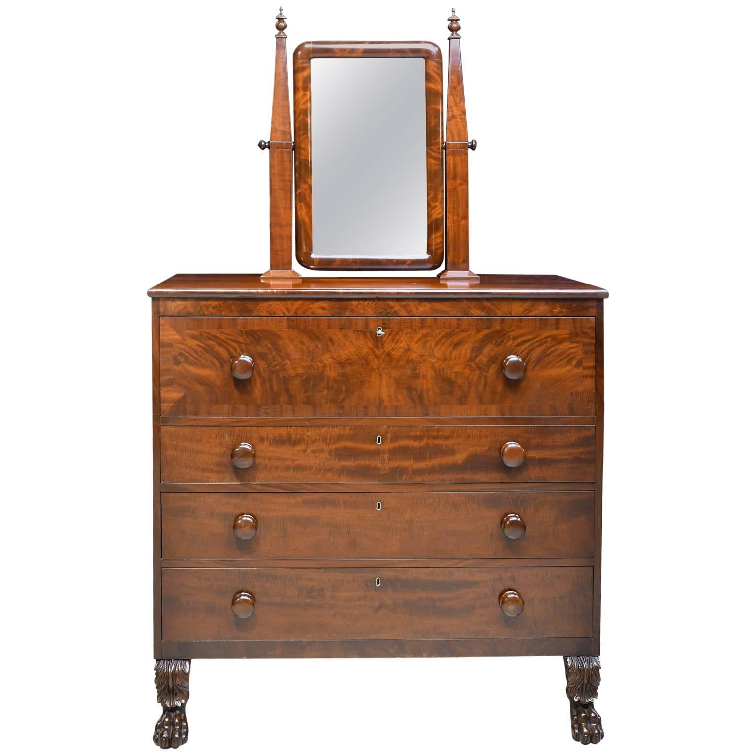 An American Empire chest in mahogany with four drawers with original turned knobs, and with original vanity mirror with square tapering columns. Chest rests on lion's paw feet with acanthus carvings on the front, and original turned back feet.