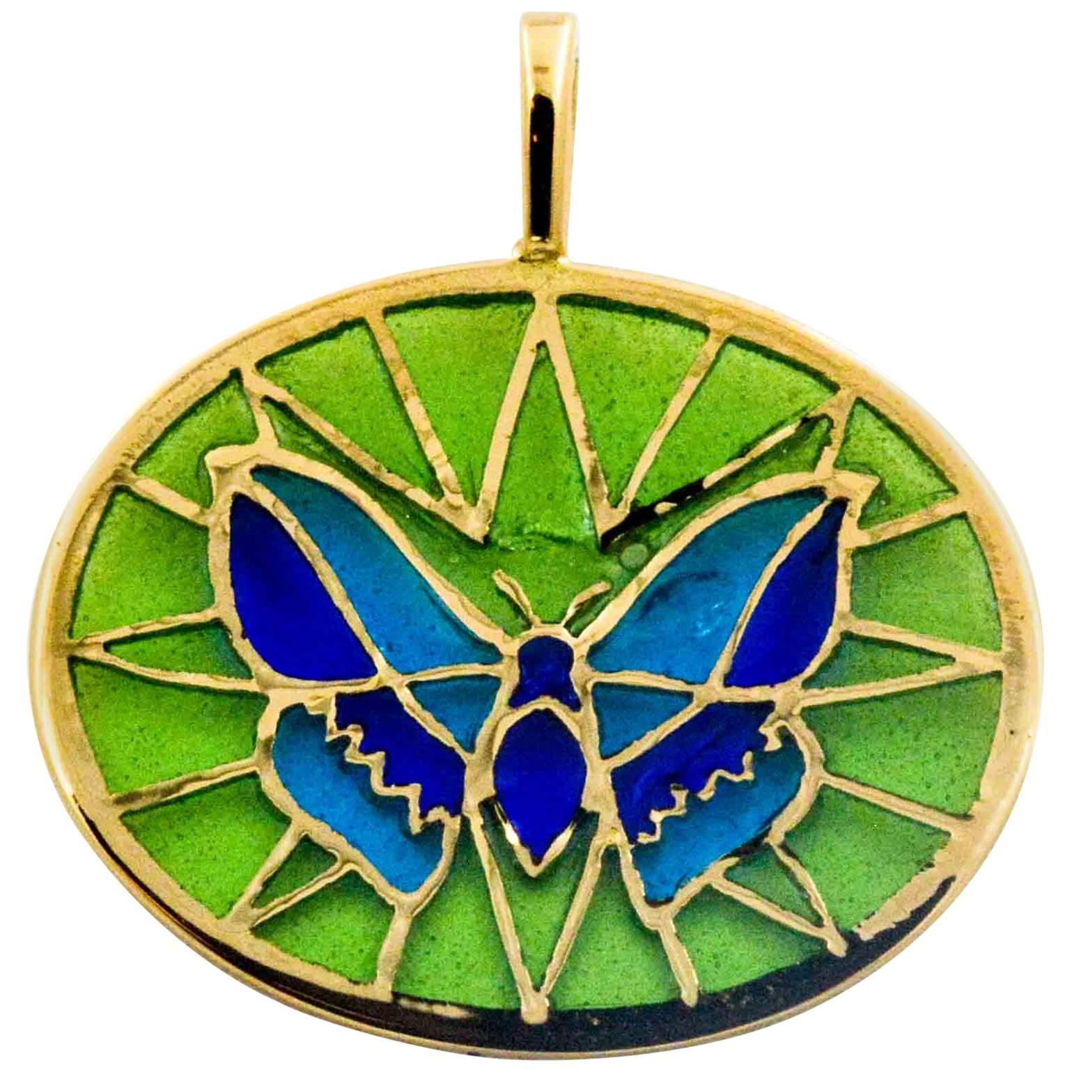 On a pale green background, a two color dark and light blue butterfly is crafted in the art of Plique A Jour glass enameling. A master craftsman created this delicate little butterfly with hand enameling blues and greens with 18 karat yellow gold as