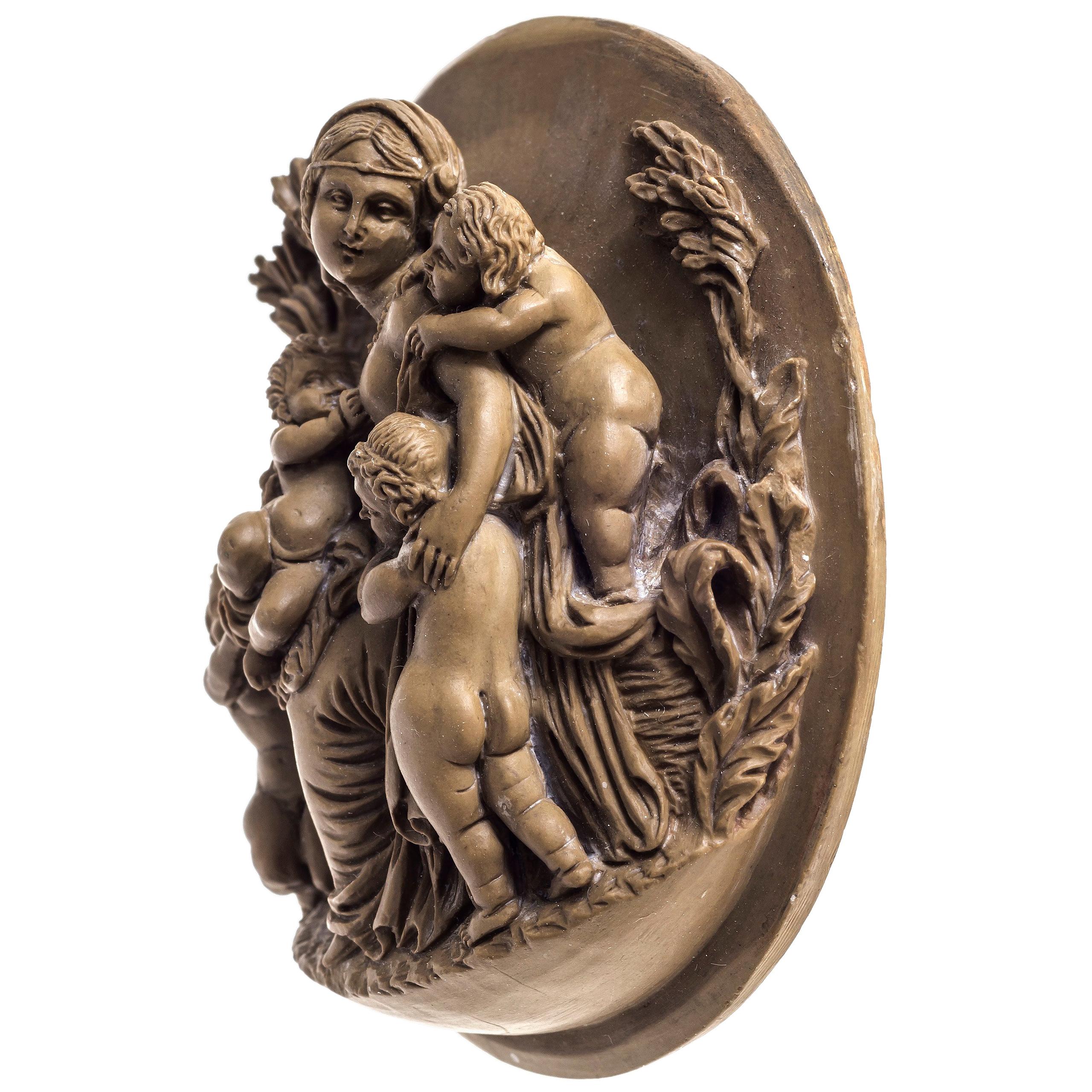 Unmounted soap stone cameo in high relief of a sitting caritas (charity) surrounded by four children in a garden. She is bare breasted, wears a richly pleated gown and a necklace. She is nurturing one of the children, two others cling to her gown.