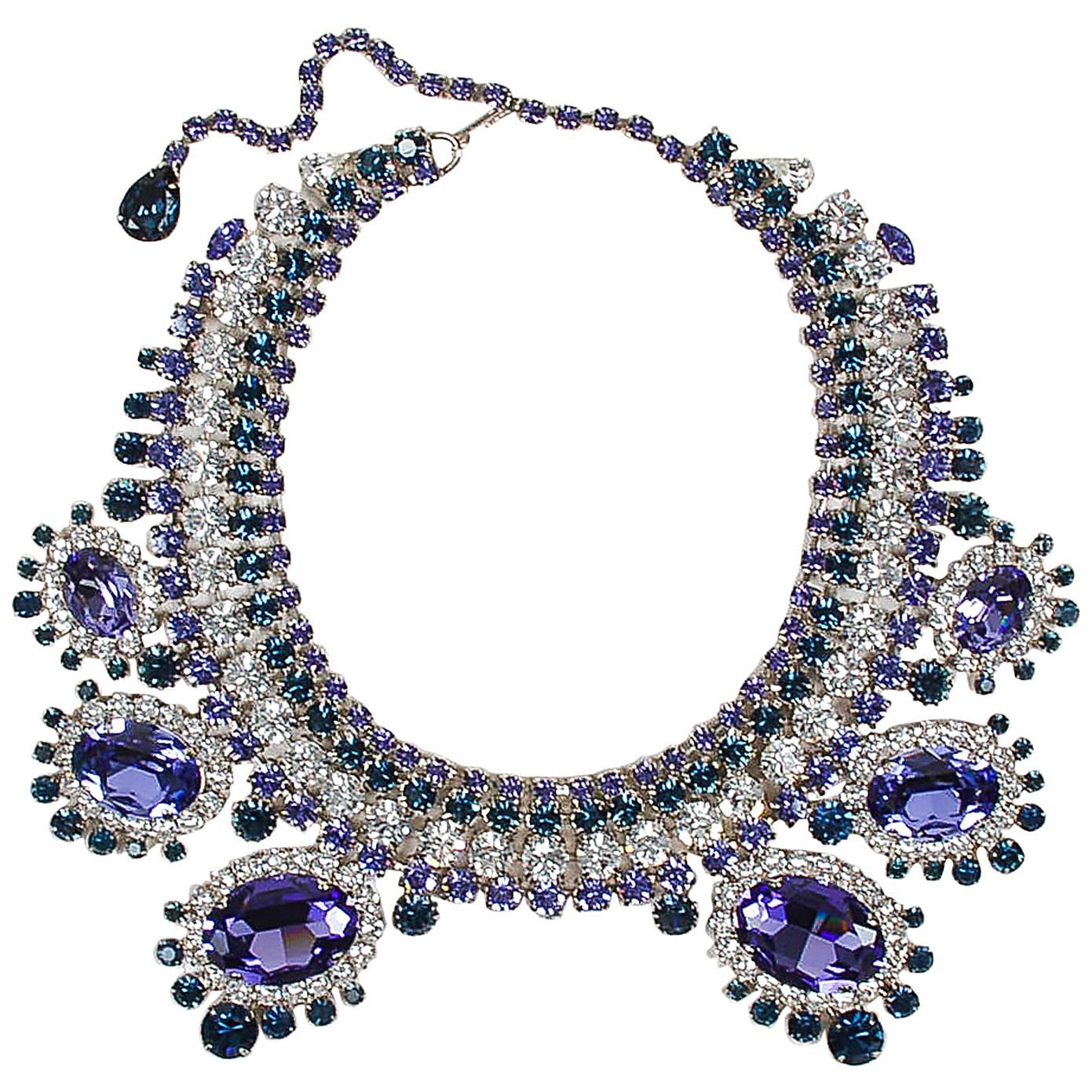 Oversized cocktail/statement bib necklace. Lustrous, light-catching rhinestone gem embellishment throughout. Purple, navy, and clear color scheme. Hook closure with extender chain. Back of necklace is engraved, 
