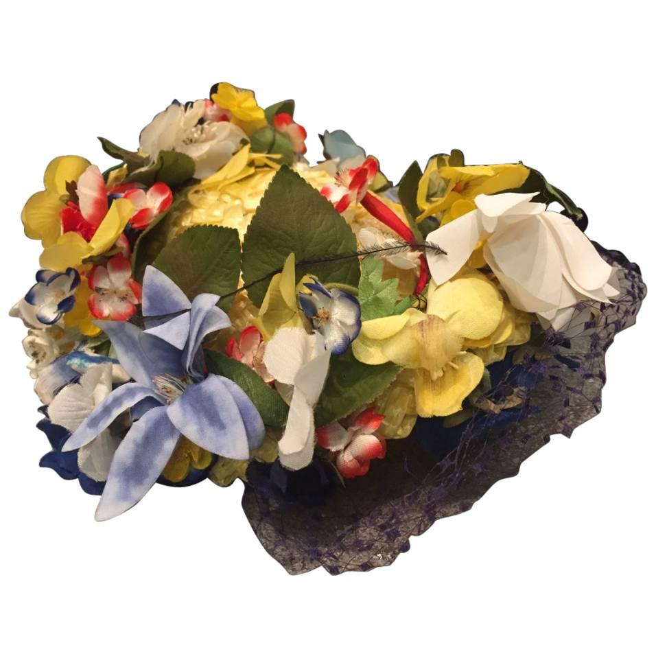 A beautiful and charming 1950s Chanda pale yellow straw hat trimmed around the small turned up brim with beautiful multi-color silk flowers and leaves.  At back is a Kelly green grosgrain bow.  A fully intact navy blue veil is attached as well as