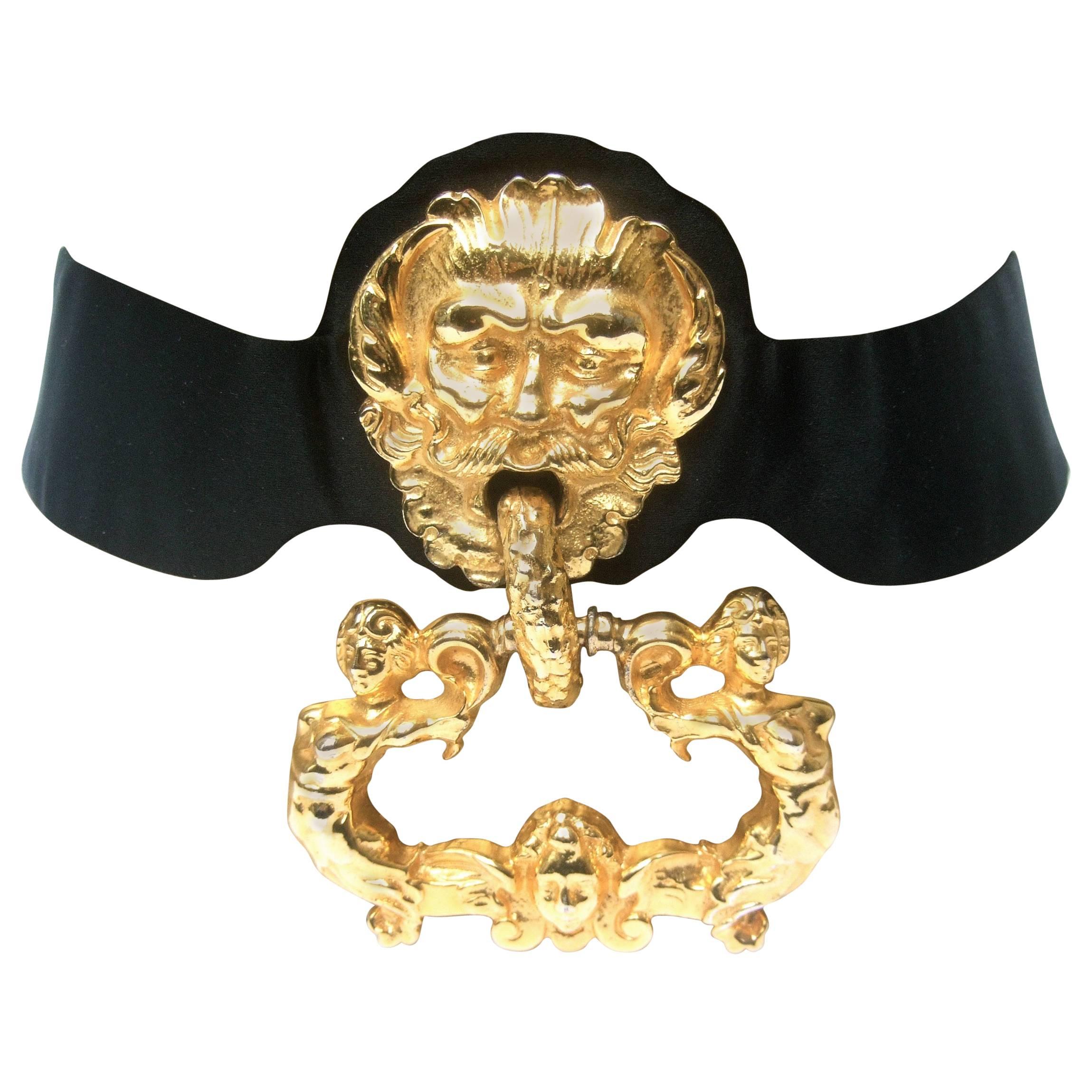 Judith Leiber Massive gilt metal door knocker satin belt c 1970s
The spectacular belt is adorned with a huge gilt metal
ornate mythical man; designed with a hinged emblem 
with a pair of ethereal woman on both sides & a cherub 
style figure at