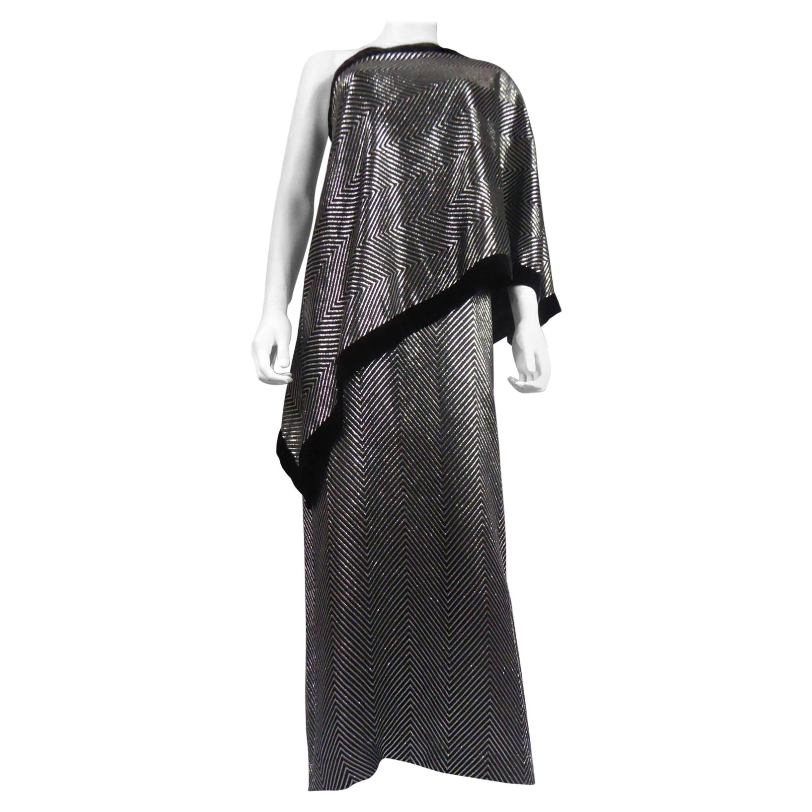Circa 1990
Italy

A Gucci velvet and silver lamé asymmetric tube dress. Wide herringbone patterns of metallic threads. Sleeve on the left side of the cape, cape point on the right side falling on the hips. White label, black graphic, G.GUCCI, size
