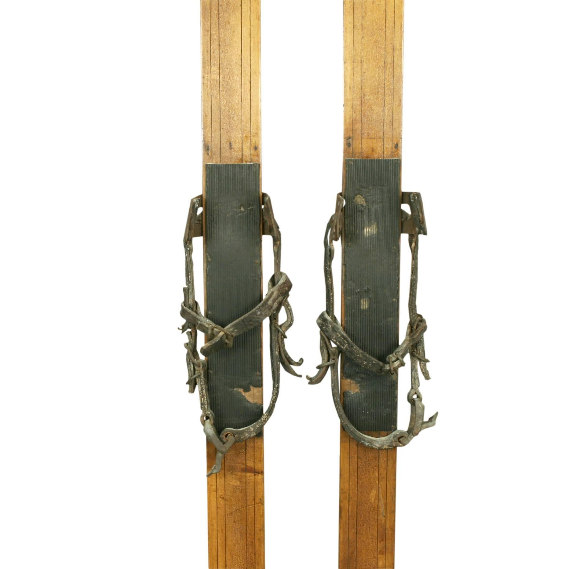 A pair of light colored ash skis with rubber footplates, galvanized toe bindings lined with leather and leather heel straps (Huitfeldt bindings). The top sides with decorative grooving and the running surfaces with a single groove. 

Ski poles are