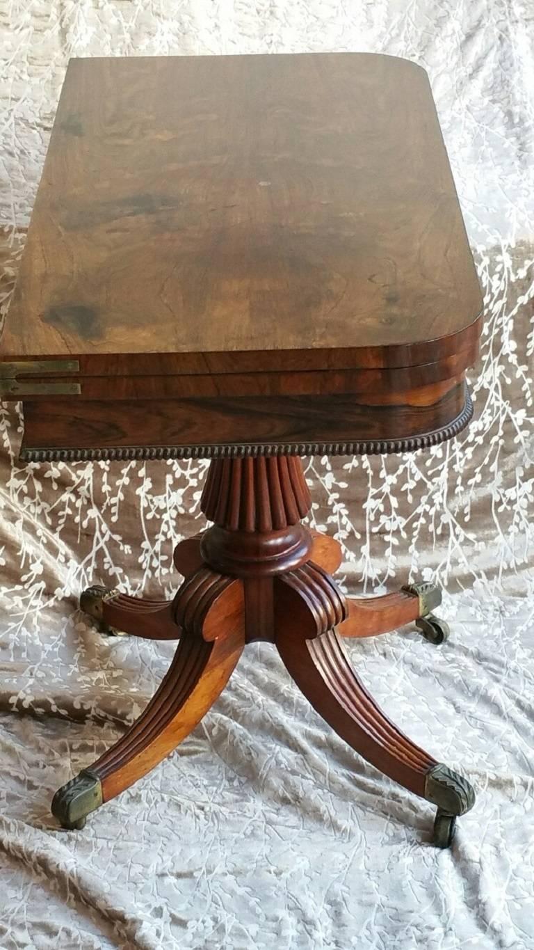 Regency rosewood card-table; the rectangular hinged and swivelling top with rounded corners, above a frieze with a split-turned beaded lower edge; on a pedestal base with a waisted and reeded baluster shaft and four hipped and reeded legs with