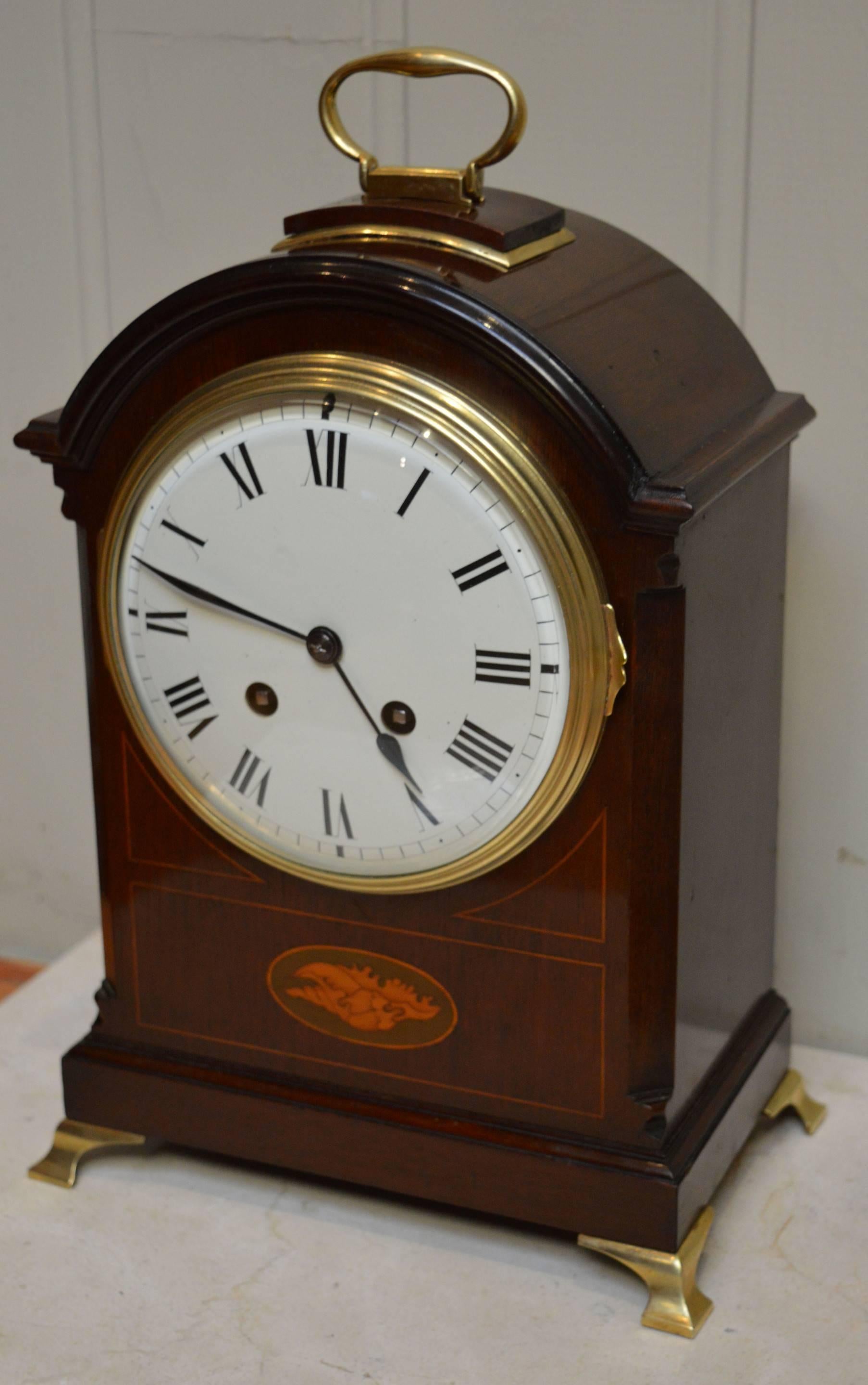 An early 20th Century solid mahogany striking bracket clock. It has a traditional break arch case, with a brass carrying handle, canted corners and brass ogee bracket feet. The front panel is inlaid with a satinwood shell and boxwood stringing. It