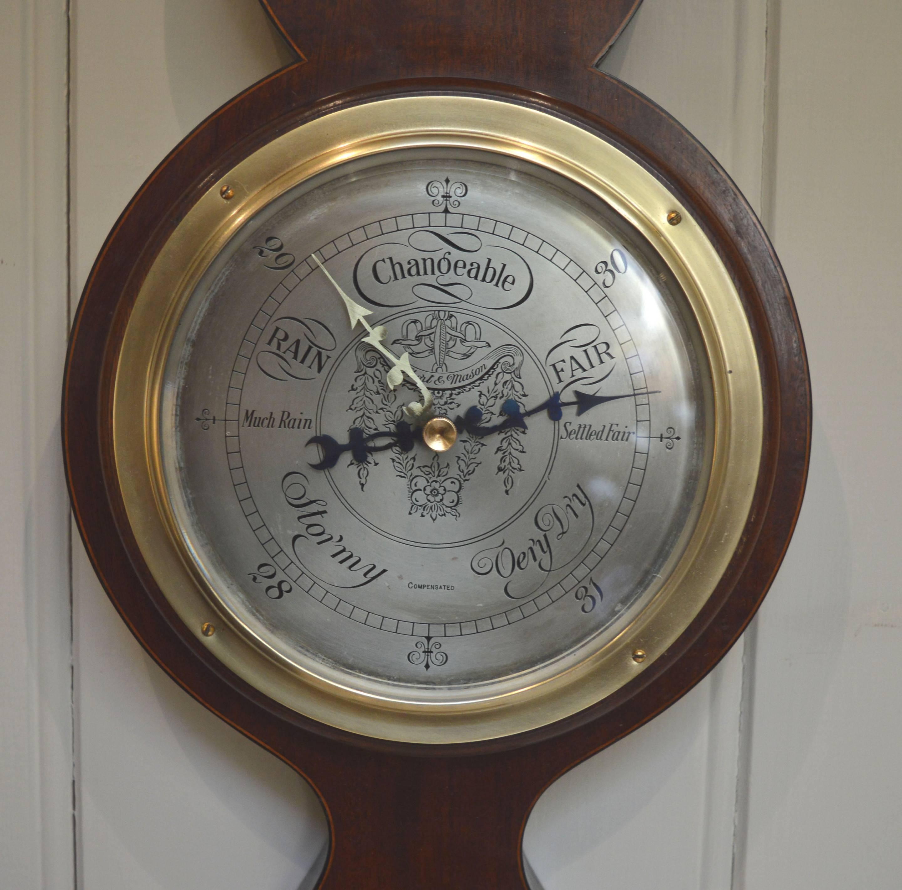 A solid mahogany android banjo barometer. Its case is inlaid with inlaid satinwood floral panels and n architectural pediment with a brass finial. It has a brass bezel and an engraved silvered dial signed by the makers Short and Mason of London. It